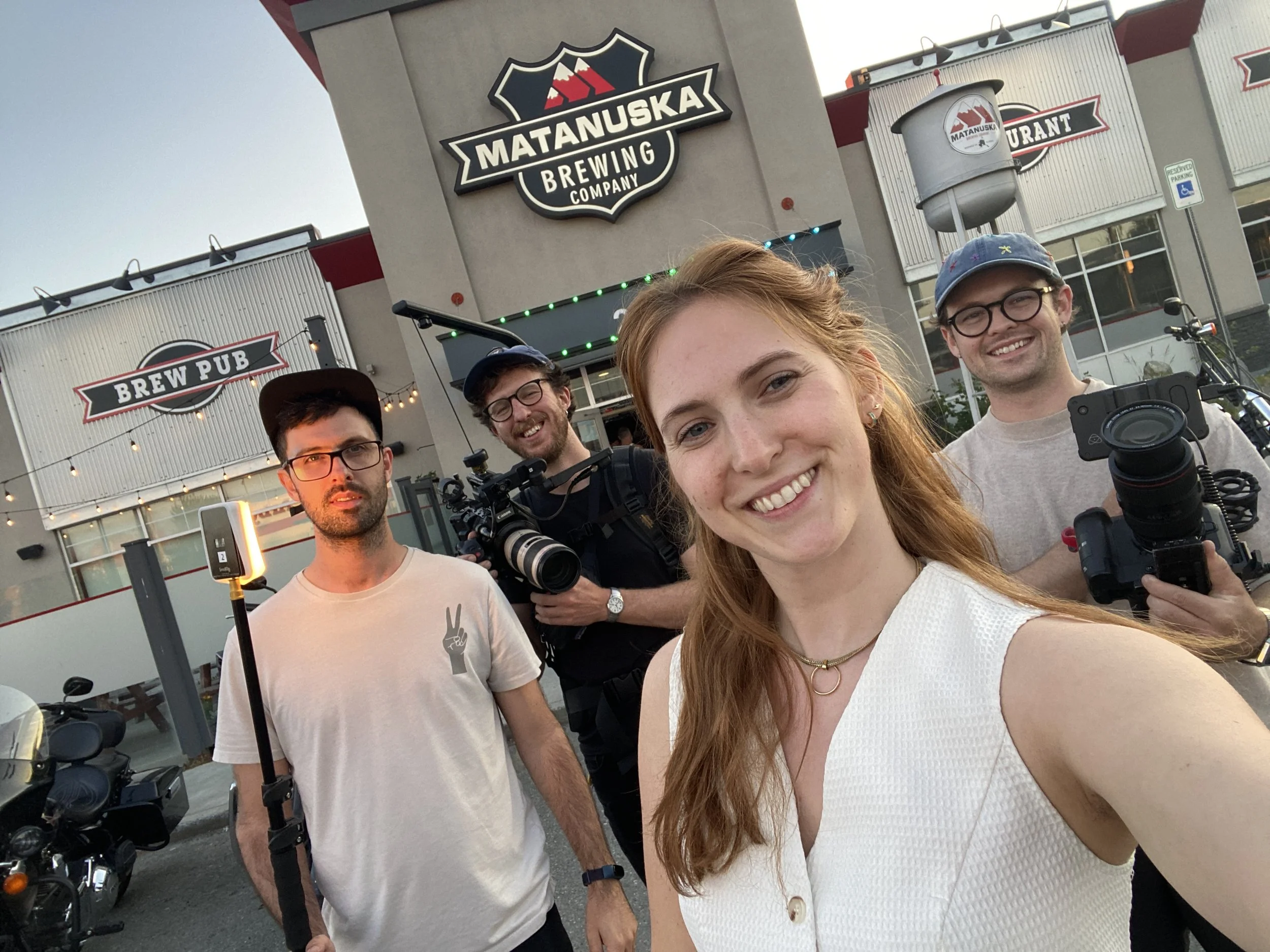 A group of five young adults standing outside Matanuska Brewing Company, some holding camera equipment, smiling for a selfie, in front of a building with signs for Brew Pub and Brewing Company.