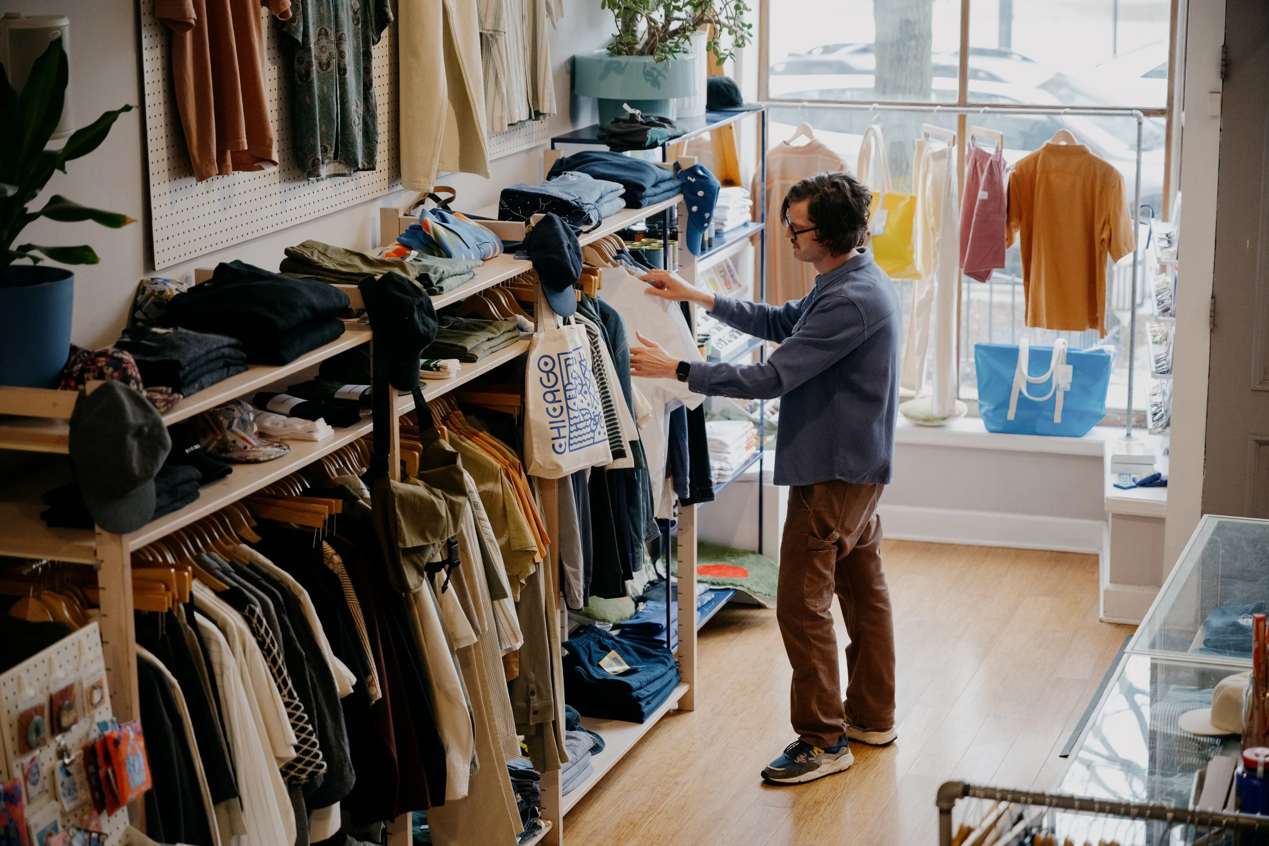 A man shopping in a clothing store, browsing through folded clothes on shelves and hanging garments. The store has large windows with hanging clothes outside and various items on display.