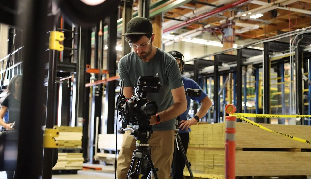 A man operating a professional video camera on a tripod inside a busy woodworking or construction workshop with people working in the background. Director of Photography, Editor, Gaffer Xander Bruce.
