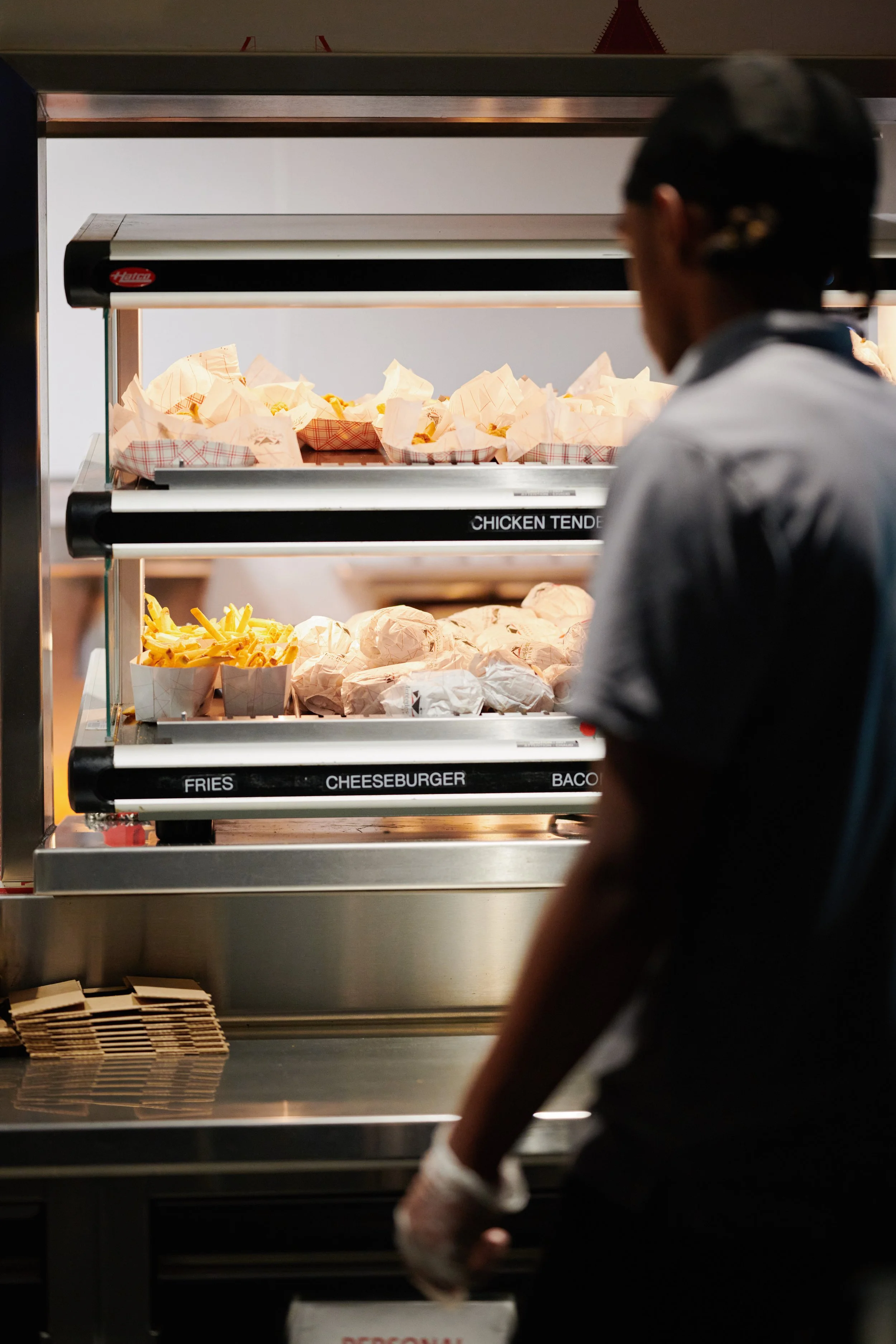 A person working at a food service station with a warming display case of fried chicken tenders, fries, cheeseburgers, and bacon, with paper food trays and wrappers. Restaurant lifestyle photography. 