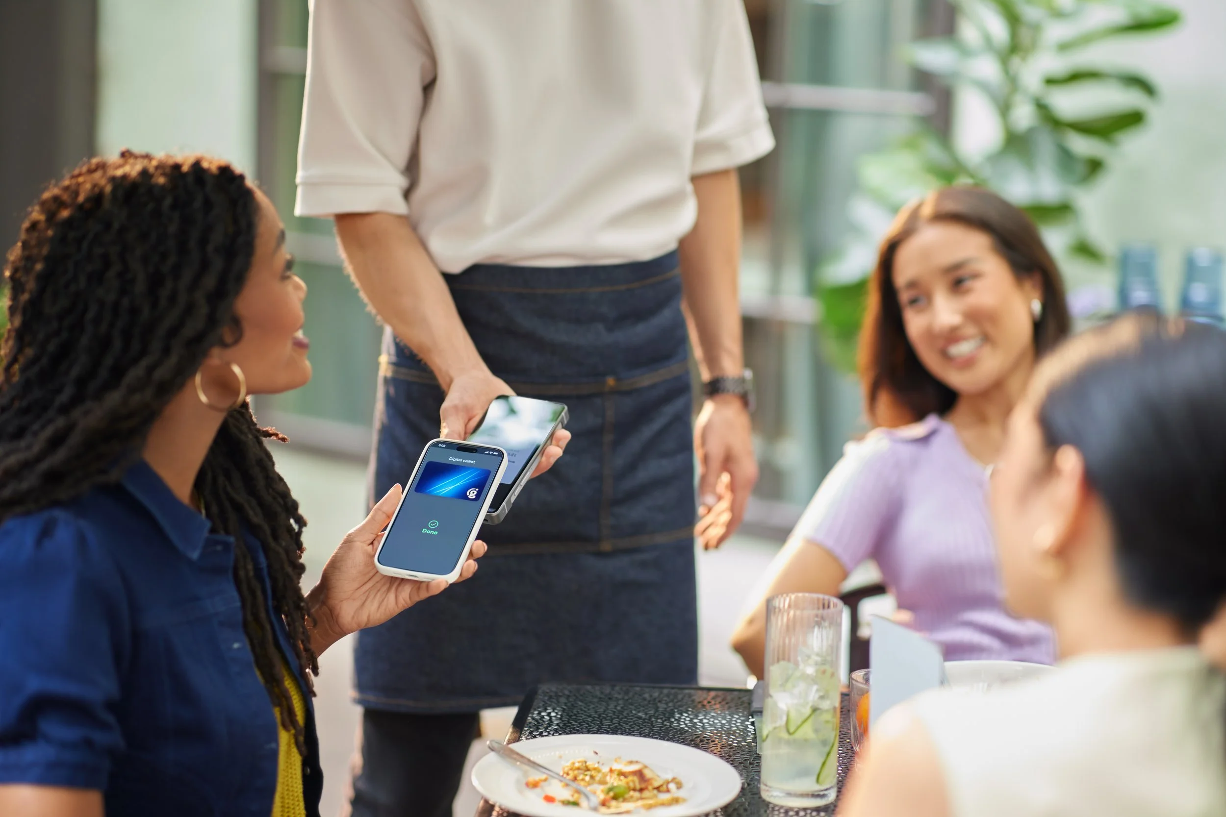 People sitting around a table with food and drinks, one person holding a smartphone, in a bright room with large windows and green plants.