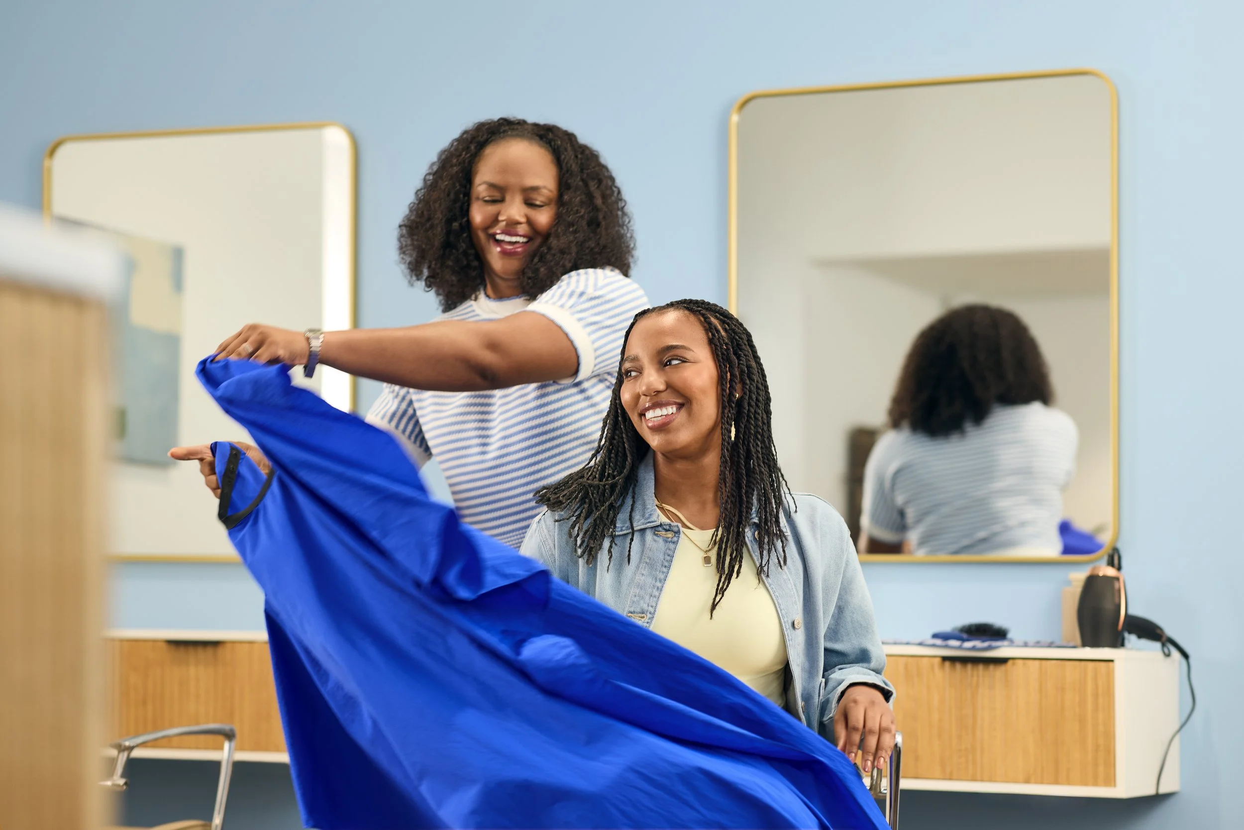 Two women with dark curly hair are near mirrors in a room with pastel blue walls, one of them is putting on a blue jacket.