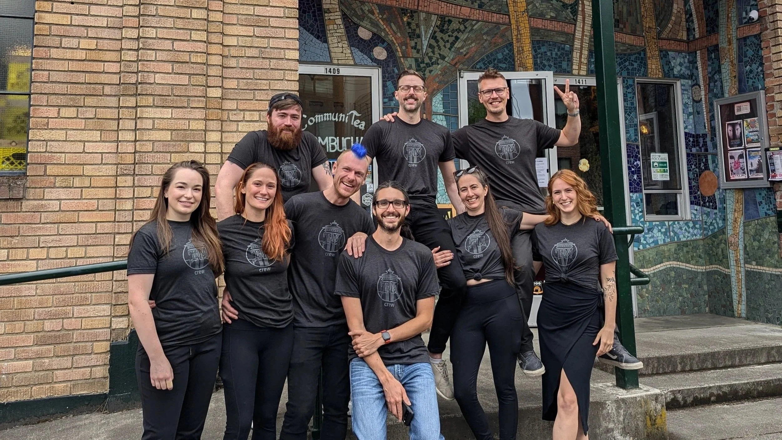 Group of nine people standing outside a building with a brick wall and colorful mosaic artwork, smiling and wearing black production crew t-shirts that represent Task Master Seattle.