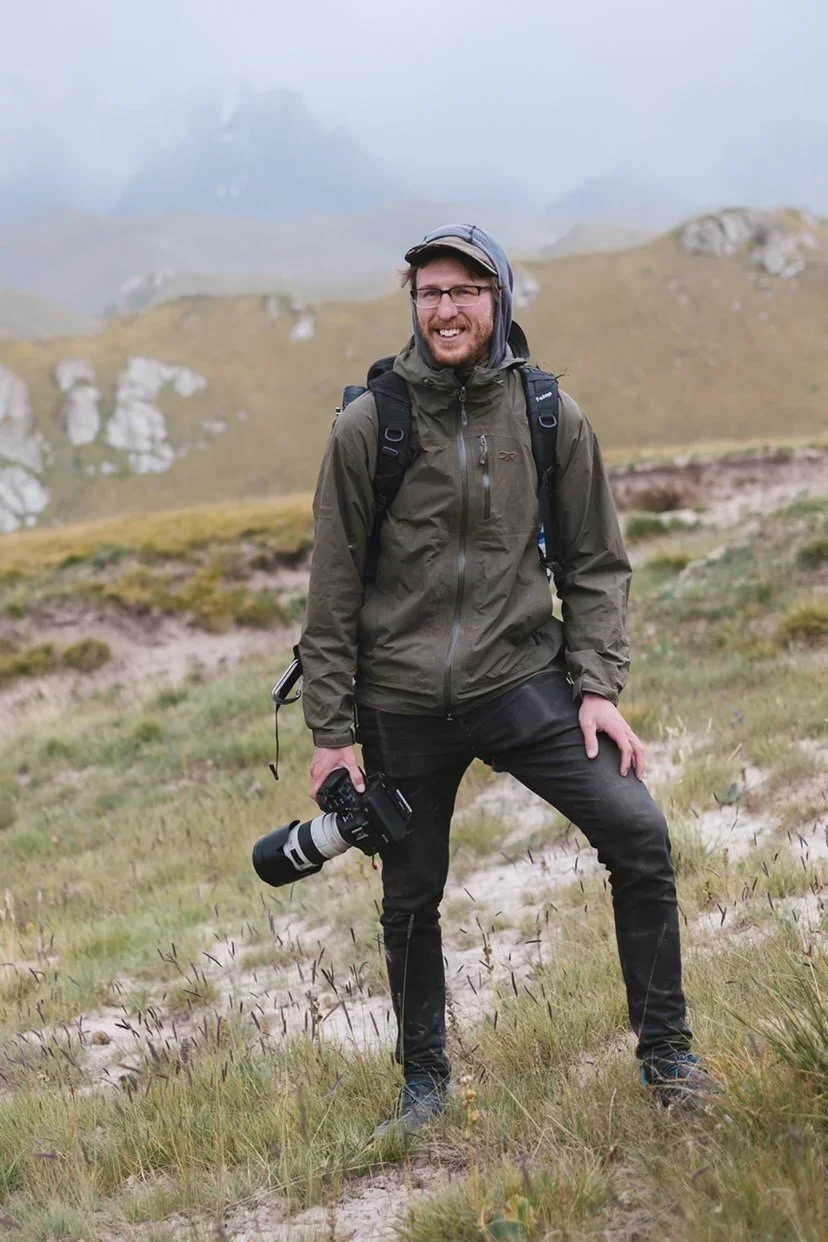 A man hiking outdoors with a camera in hand, wearing a brown jacket, black pants, glasses, and a backpack, standing in a grassy and rocky landscape with hills and cloudy sky in the background. Director of Photography, Editor Brady Lawrence.