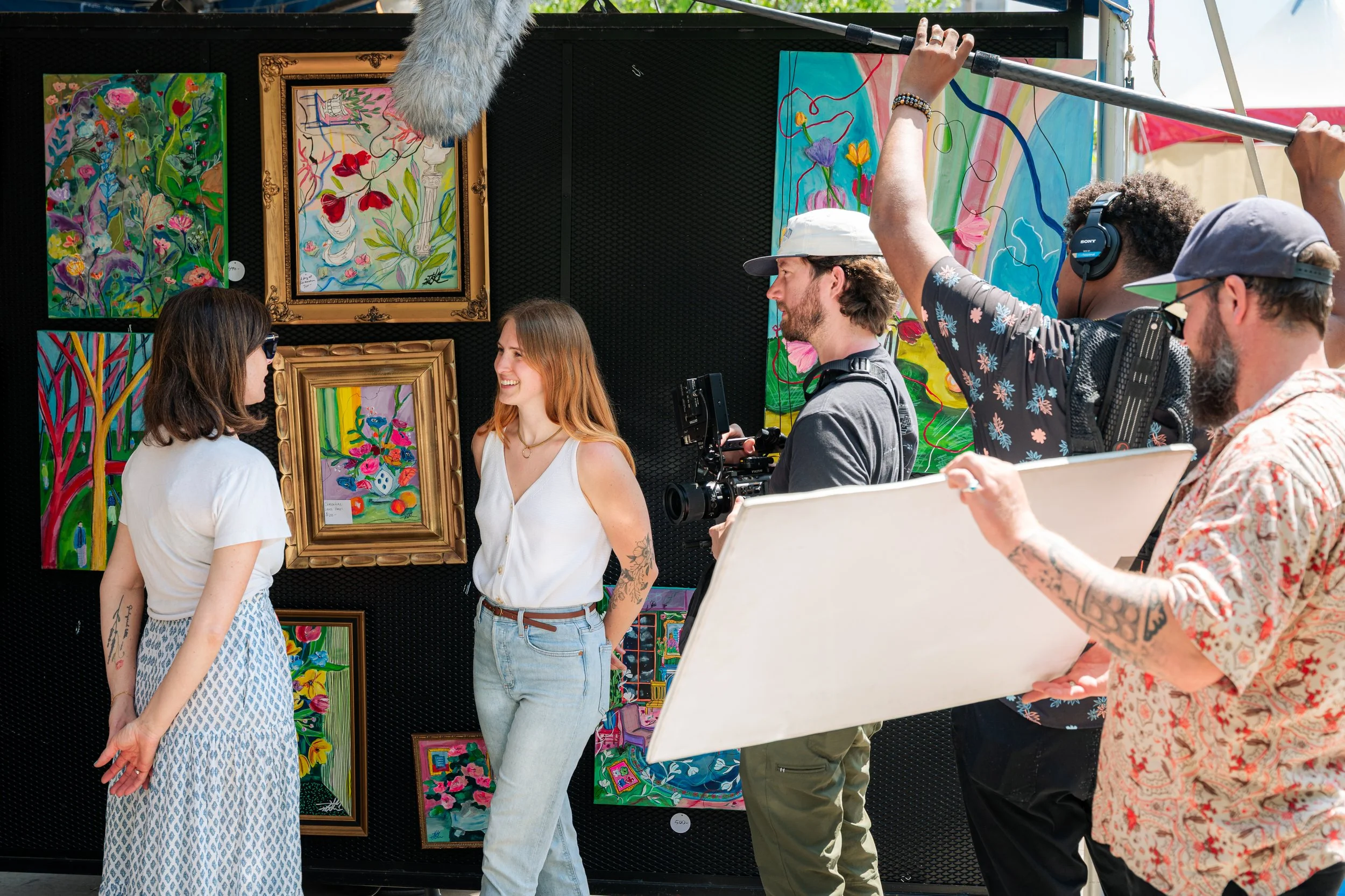 Four women and two men standing and engaging at an outdoor art exhibition, with colorful paintings displayed on black walls behind them. The women are facing each other, smiling, with one in a white top and yellow bracelet, and the other in a patterned dress. One man is holding a camera, and another man is holding a gray easel and wearing headphones, all surrounded by vibrant artwork.