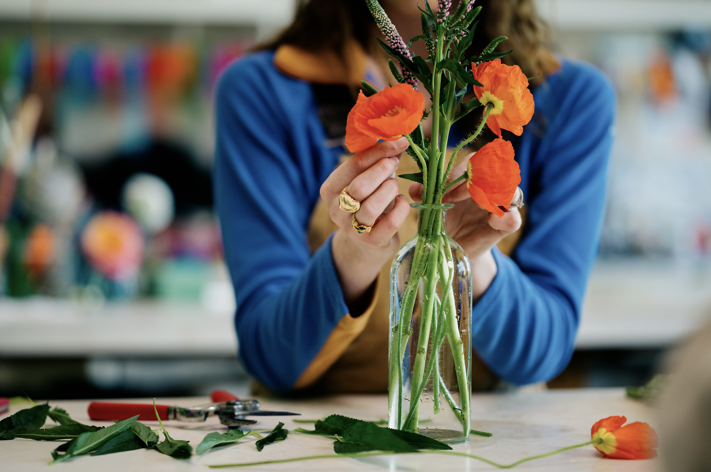 Person arranging orange and purple flowers in a glass vase at a flower arranging workshop. Retail and lifestyle photography featuring bold colors. Detail oriented photography that highlights the small moments of a story.
