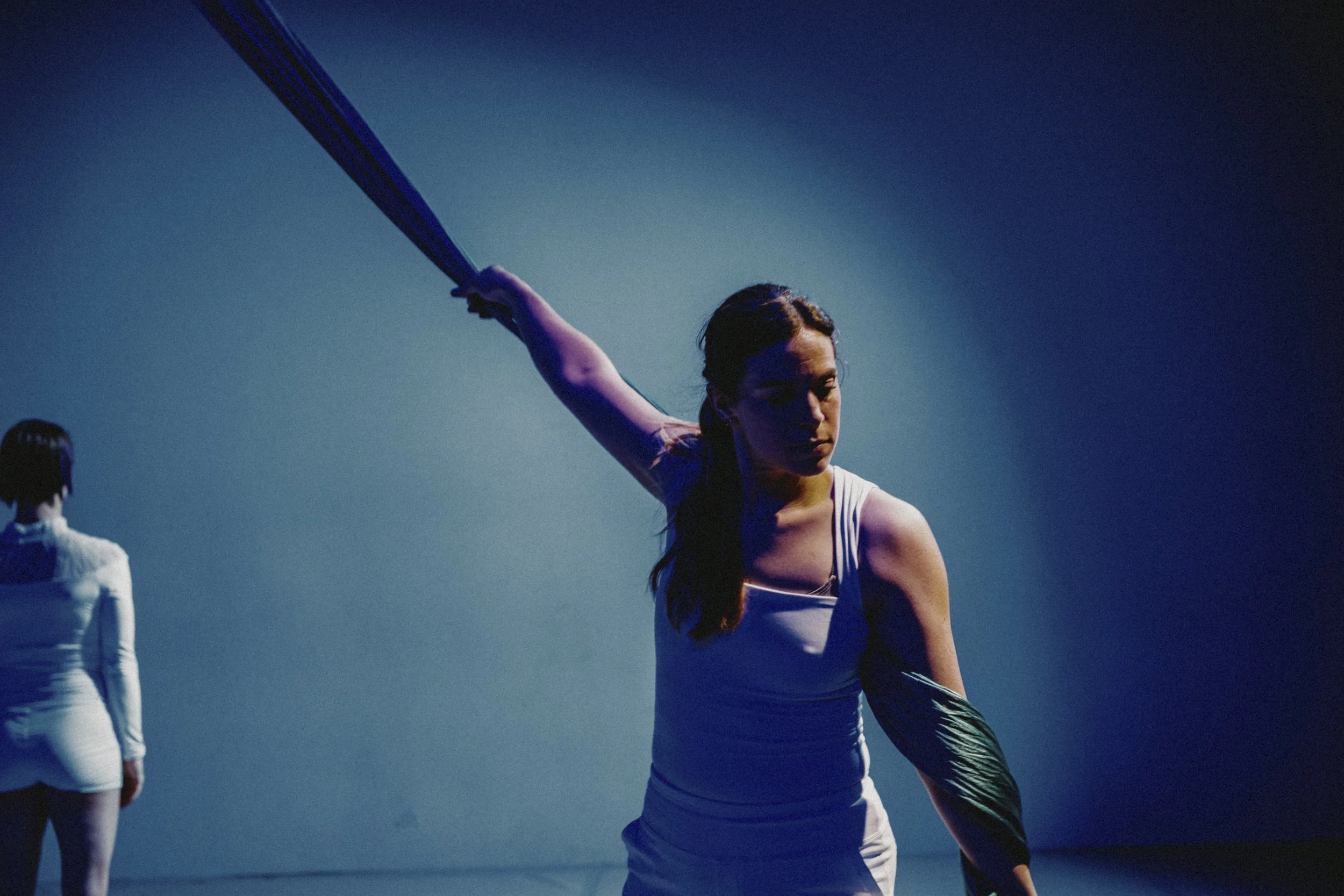 A young woman in athletic attire performing a dance or exercise move, holding what appears to be a baton or similar object, with two other women in the background against a dark blue background.