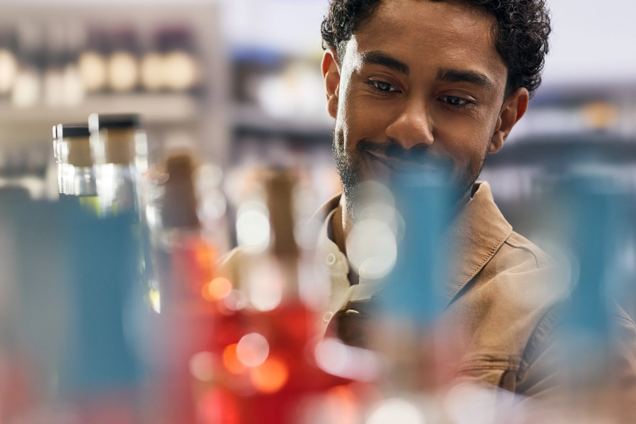 A young man with curly dark hair and a beard, smiling, looking at colorful objects on a store shelf, with a blurred background.
