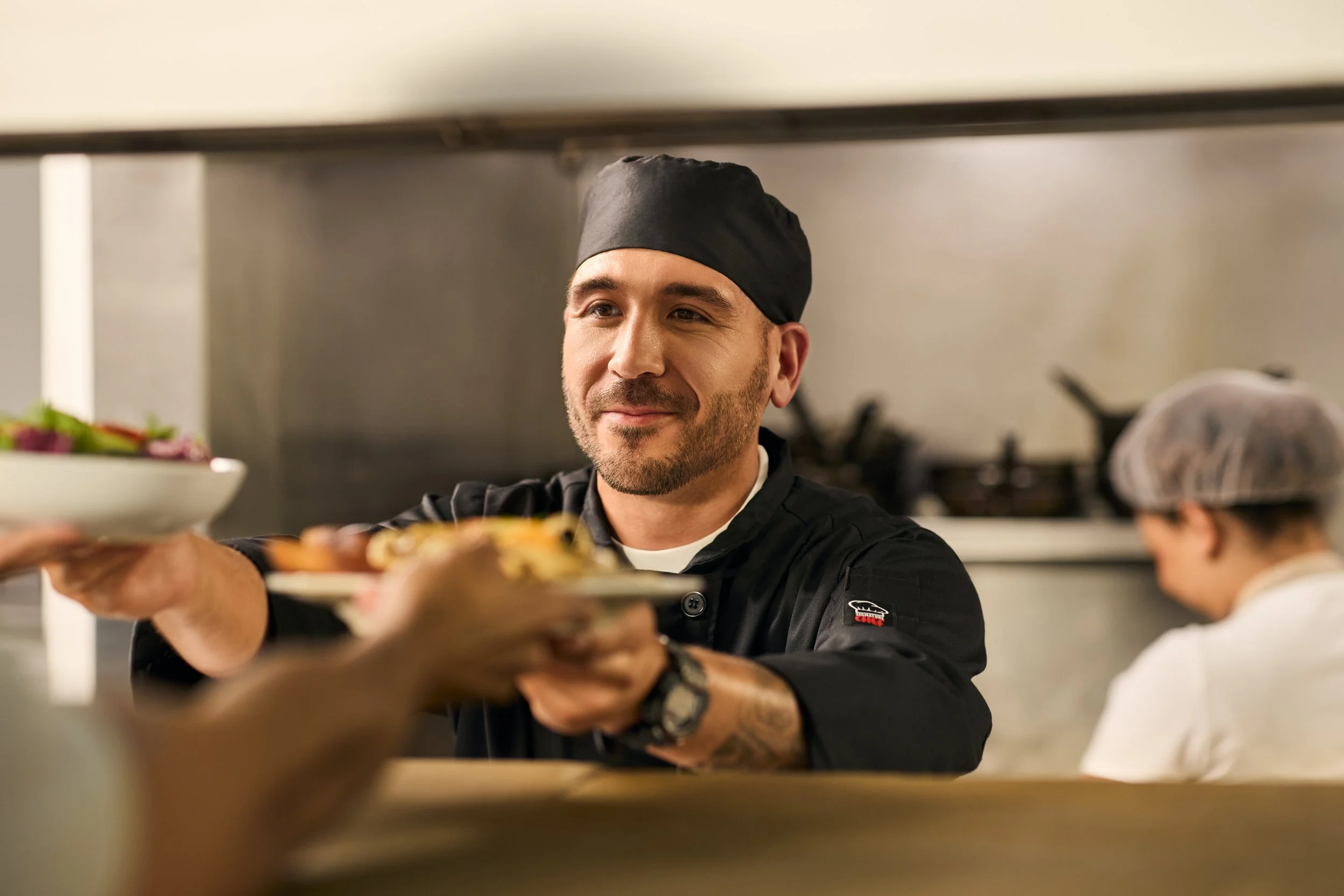 A chef in a professional kitchen offers a plate of food towards the camera.