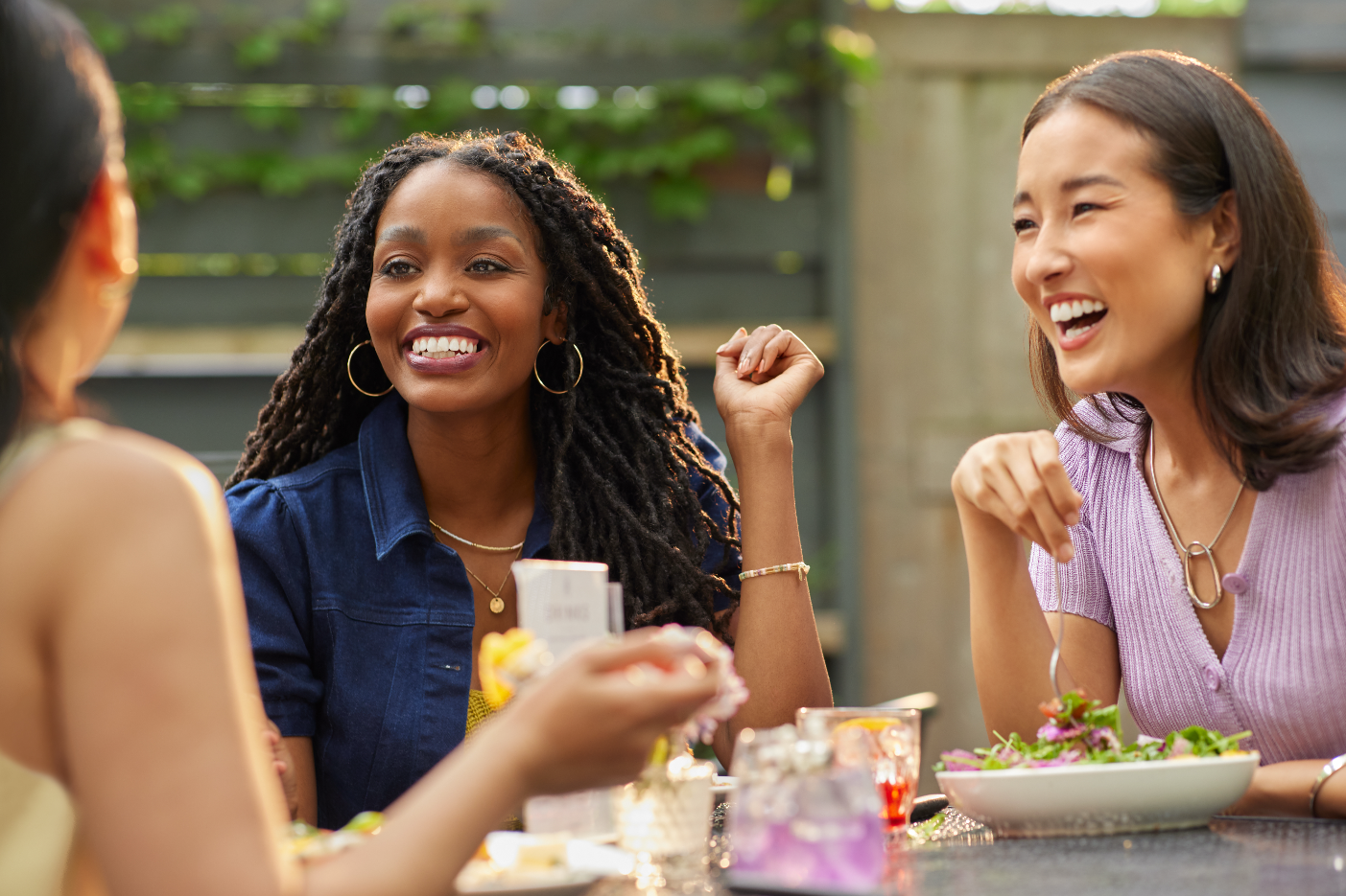Three women talking and laughing at a table outdoors, with food and drinks, during daytime. Restaurant and lifestyle photography featuring warm, bright expressions and a light and airy color grade. 