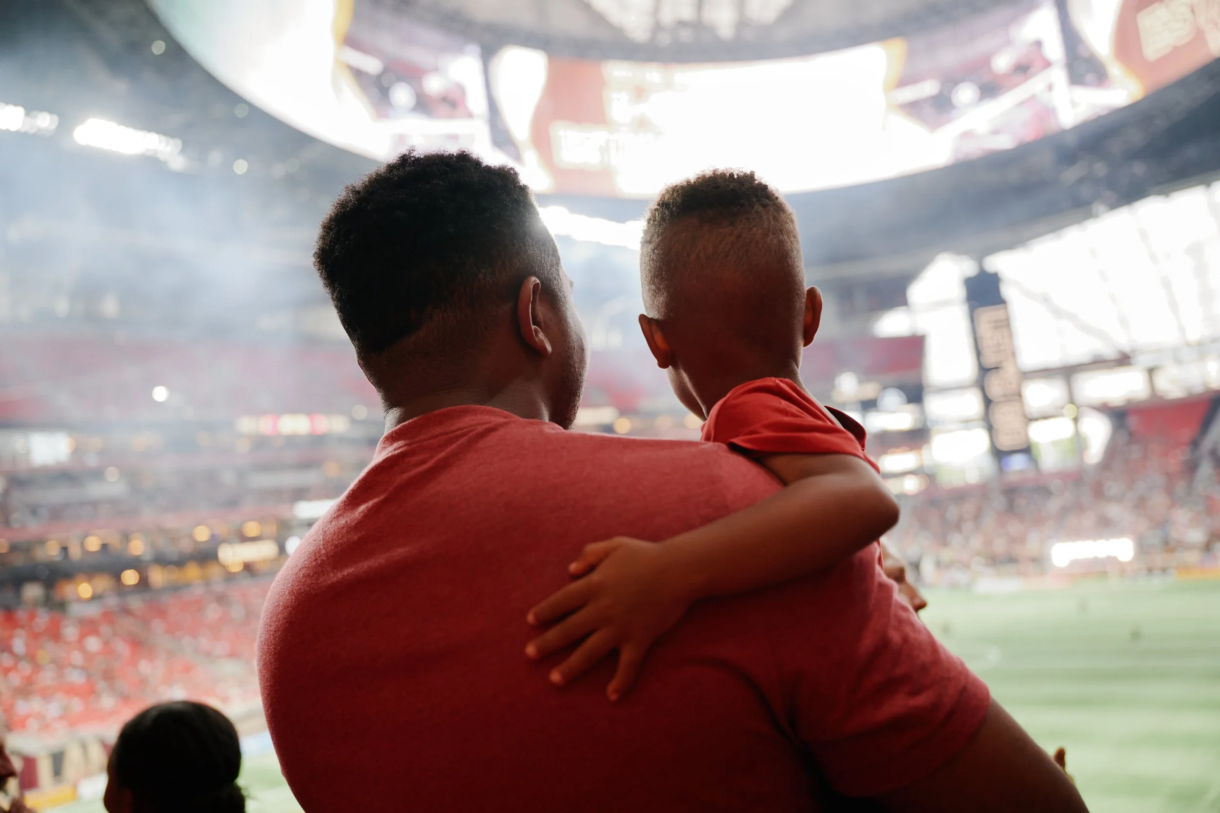 A man and his child stand together in a stadium, looking toward the field, with the child's arm around the father's shoulder, wearing red shirts, in a bright stadium filled with spectators. Sports experience photography. 
