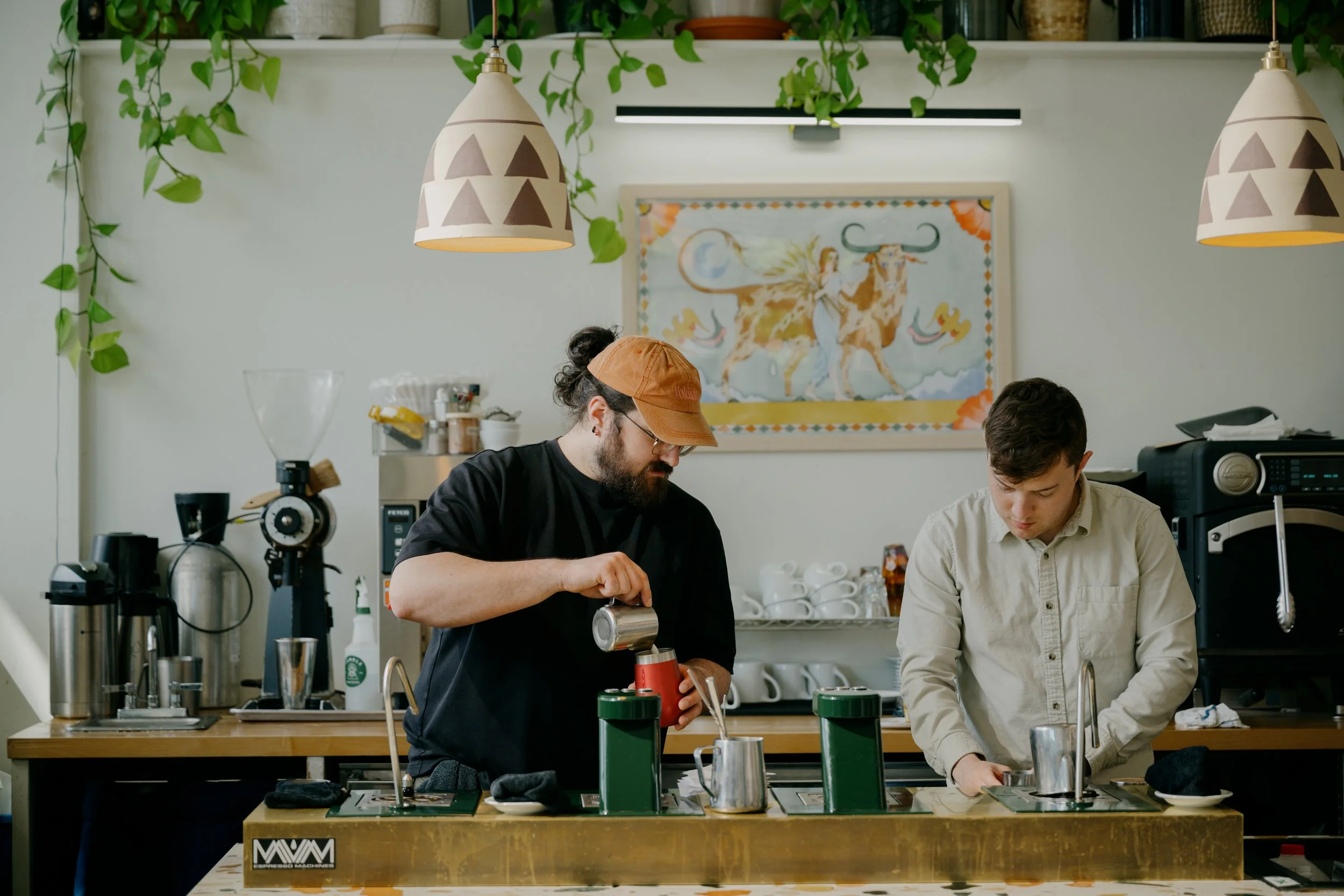Two men preparing coffee behind a counter in a cafe, with coffee-making equipment and cups, and a vibrant wall hanging depicting a woman with wings and a bull, surrounded by hanging plants and modern light fixtures.