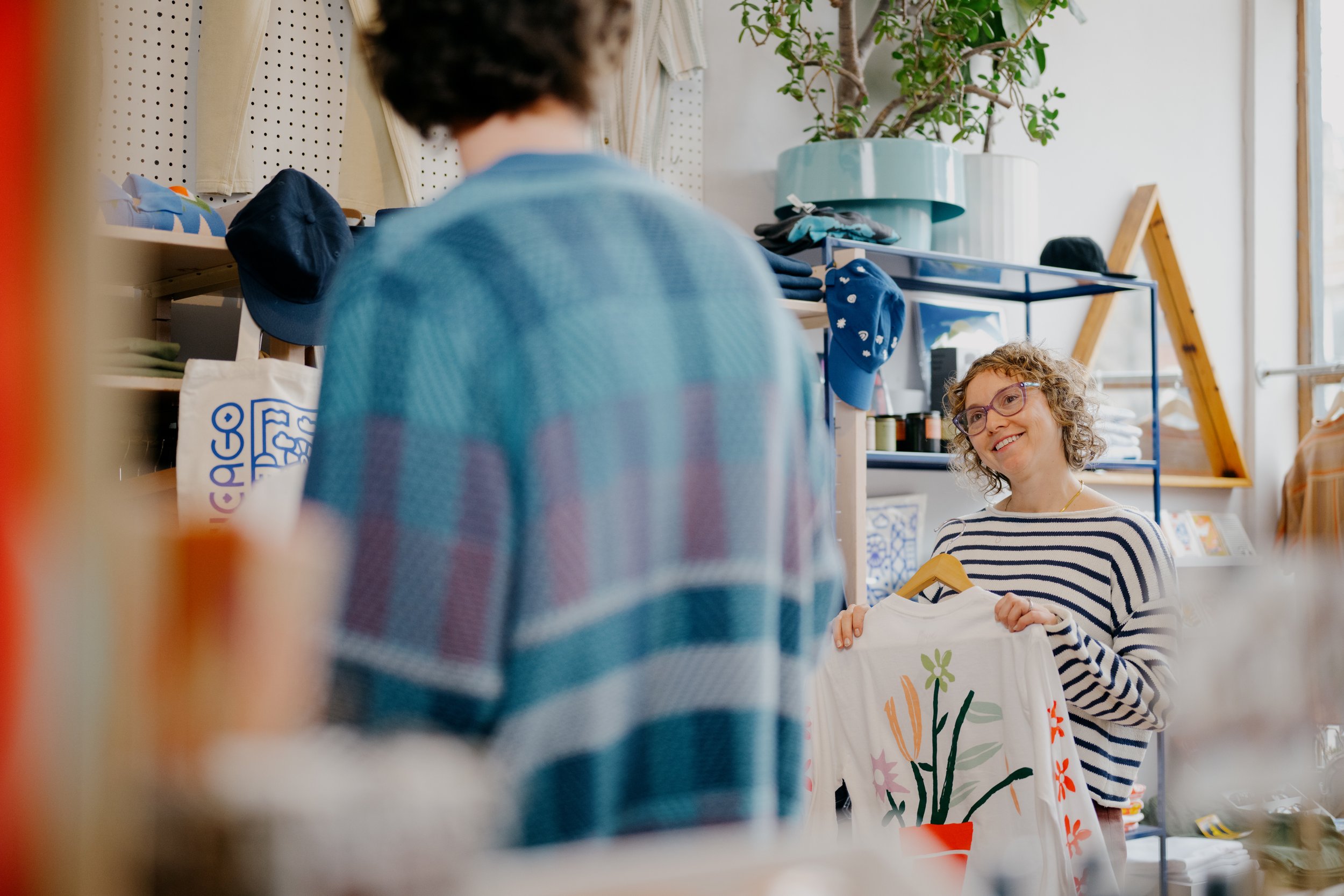 A woman with curly hair and glasses smiling and holding a white cloth with colorful flower and plant paintings in a store or market. Retail and lifestyle photography featuring warm, bright expressions and a light and airy color grade. 