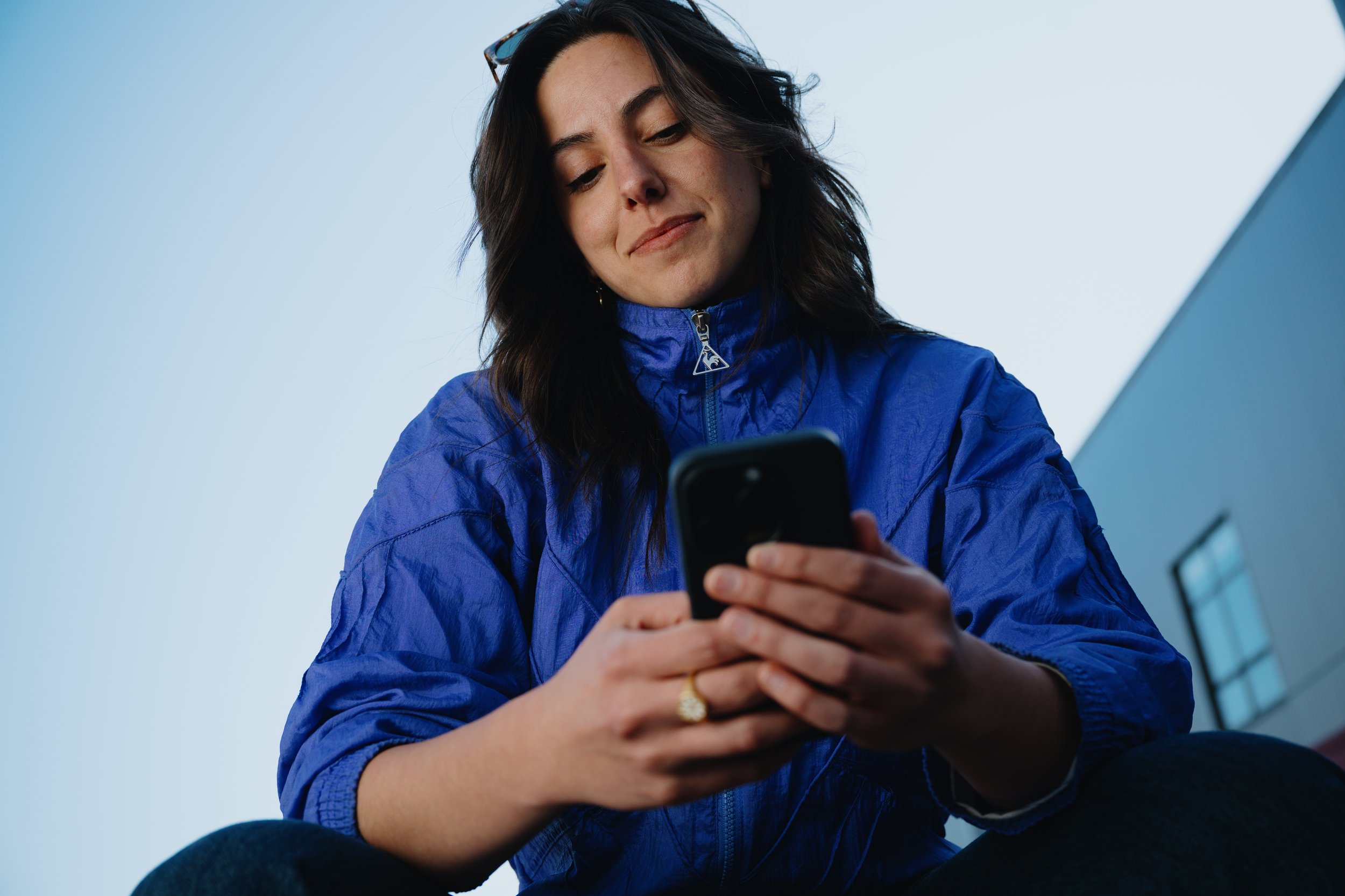 A young woman with long, dark hair and glasses resting on her head, wearing a blue jacket, looks down at her smartphone with a slight smile against a light blue sky background.