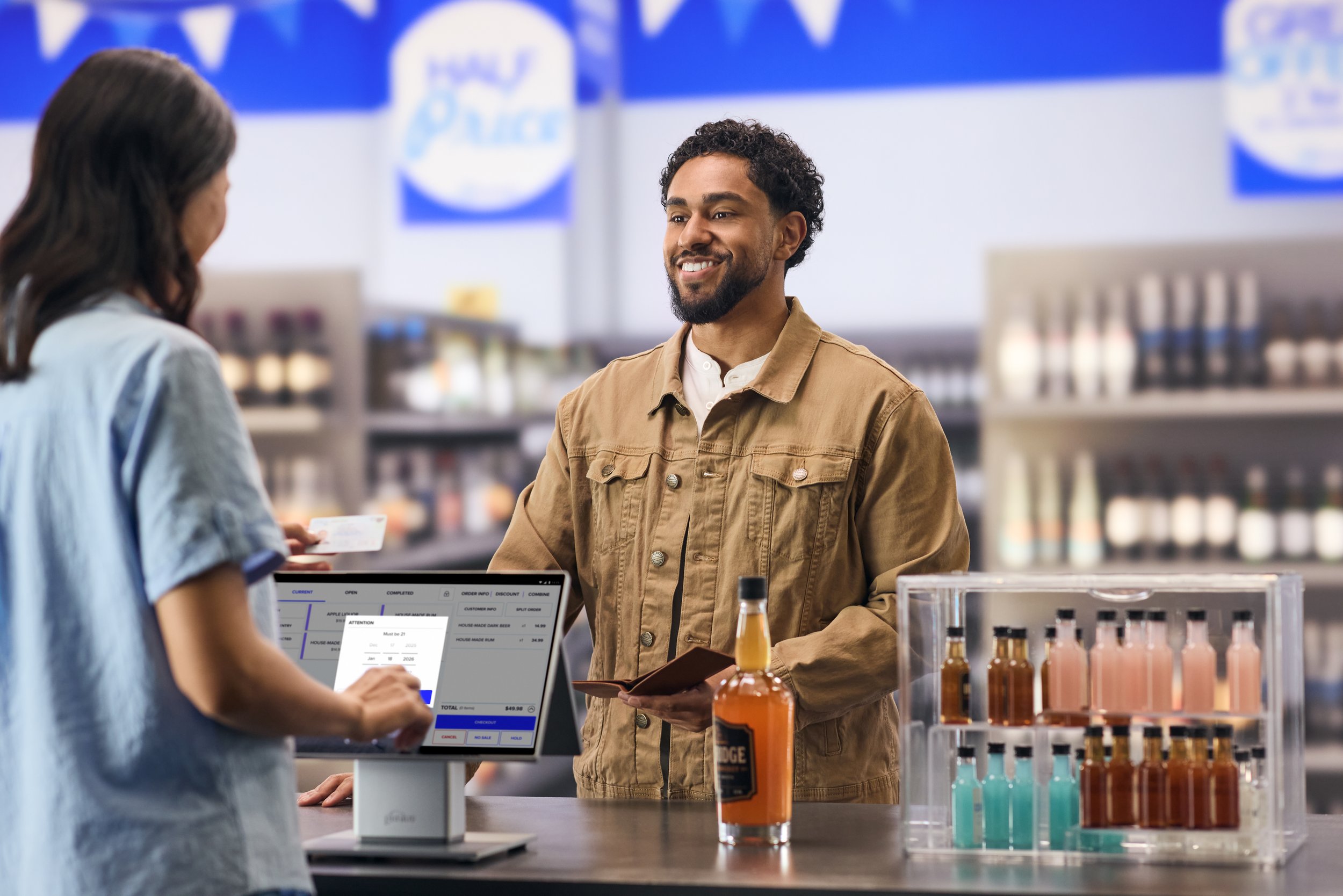 A man with curly hair and a beard smiling at a woman with long dark hair, in a store checkout counter with bottles of soda and a digital payment screen in the foreground.