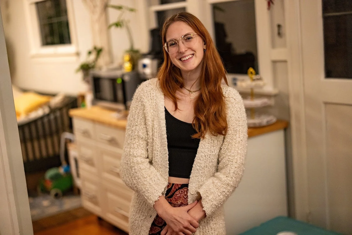 A young woman with long red hair, glasses, and a warm smile standing in a cozy kitchen, wearing a beige cardigan over a black top and patterned pants. Producer, director, 1st AD Victoria Fouke.