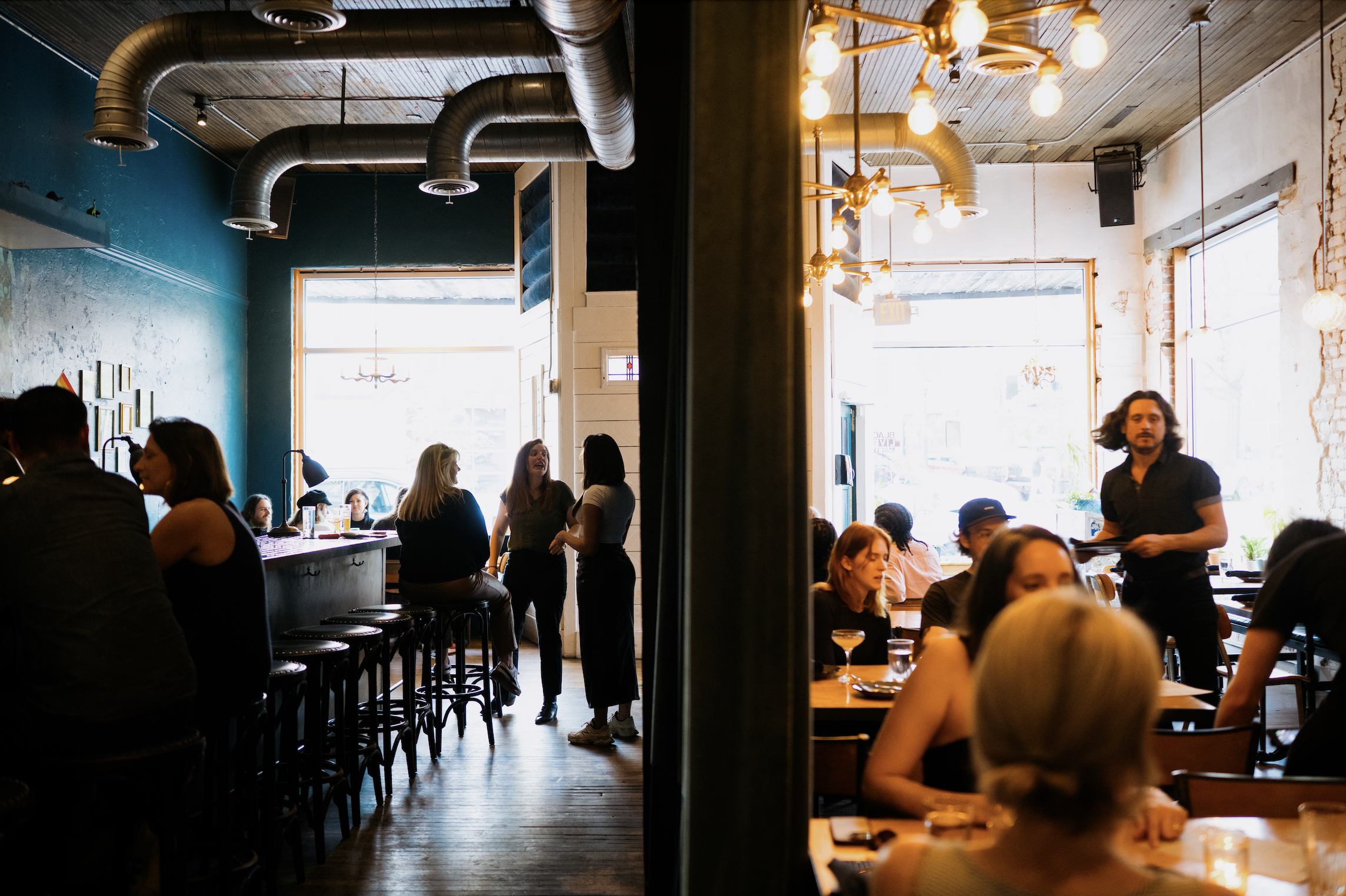 A busy restaurant interior filled with people dining and socializing, with large windows, industrial-style lighting, and exposed ductwork. Detail oriented photography that highlights the big moments of a story.