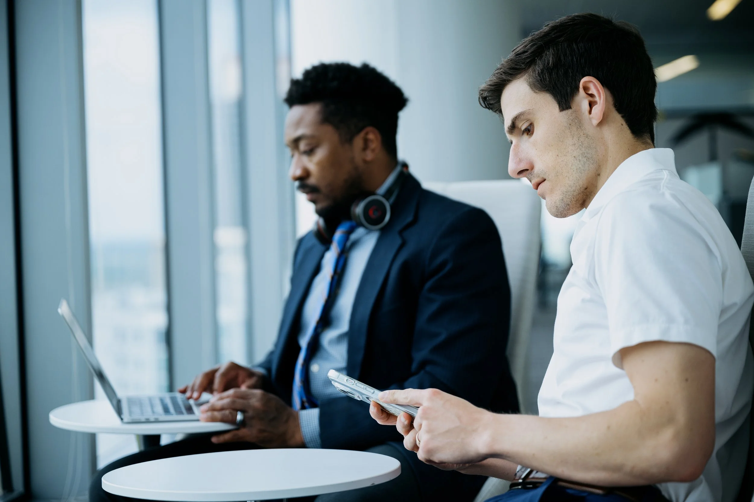 Two men sitting at white tables in a modern office or airport, one using a laptop and the other looking at a smartphone, with large windows in the background.