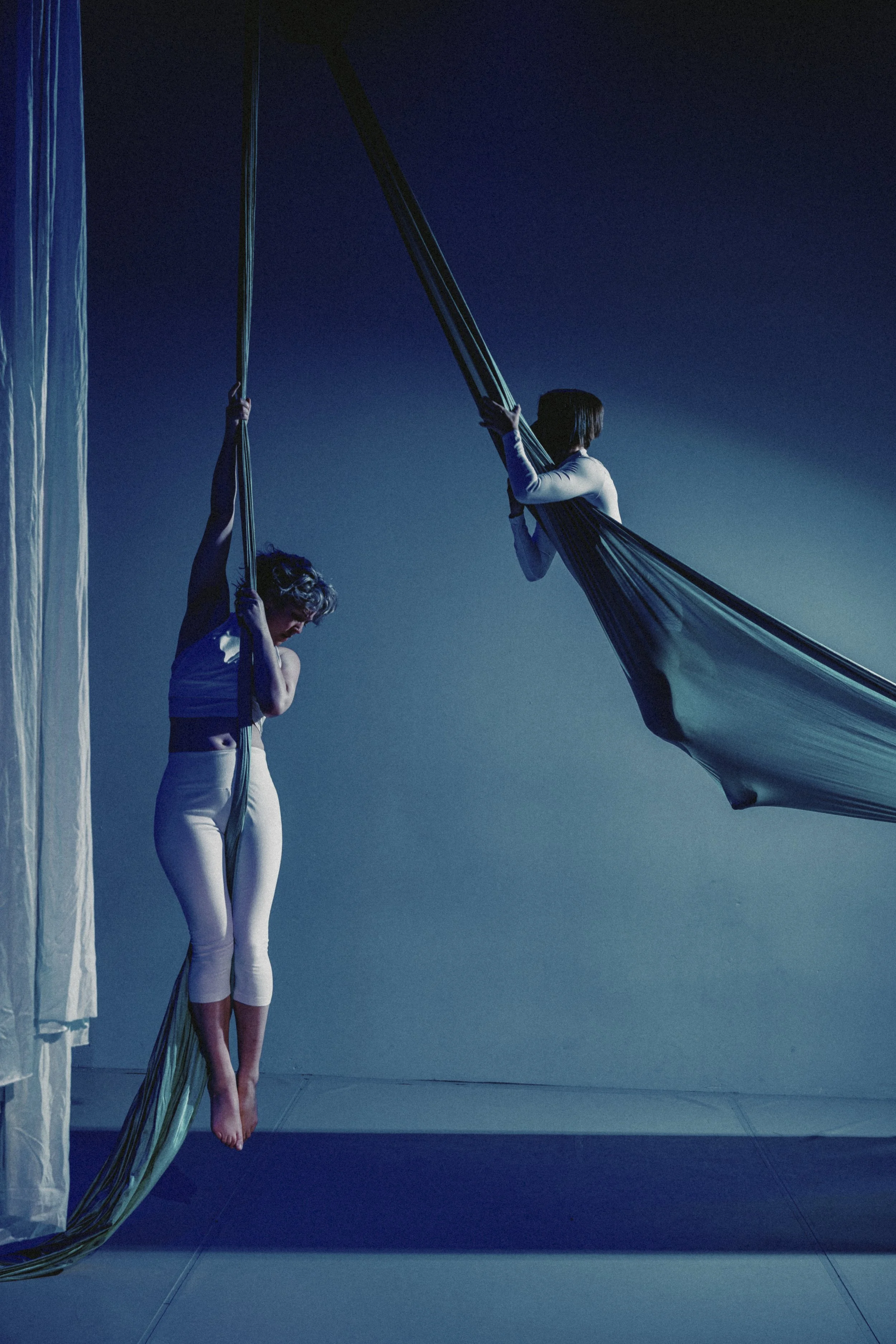 Two women in athletic wear perform aerial silk exercises on blue fabric suspended from the ceiling against a dark background. One woman stands with her eyes closed, while the other is holding onto the fabric, looking away.