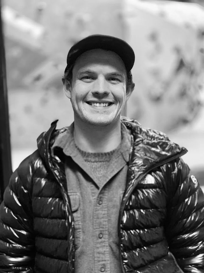 A smiling man wearing a black cap, a puffer jacket, and a gray button-up shirt in a black-and-white photo. Director of Photography, editor, Gaffer, Grip Keith Oler.