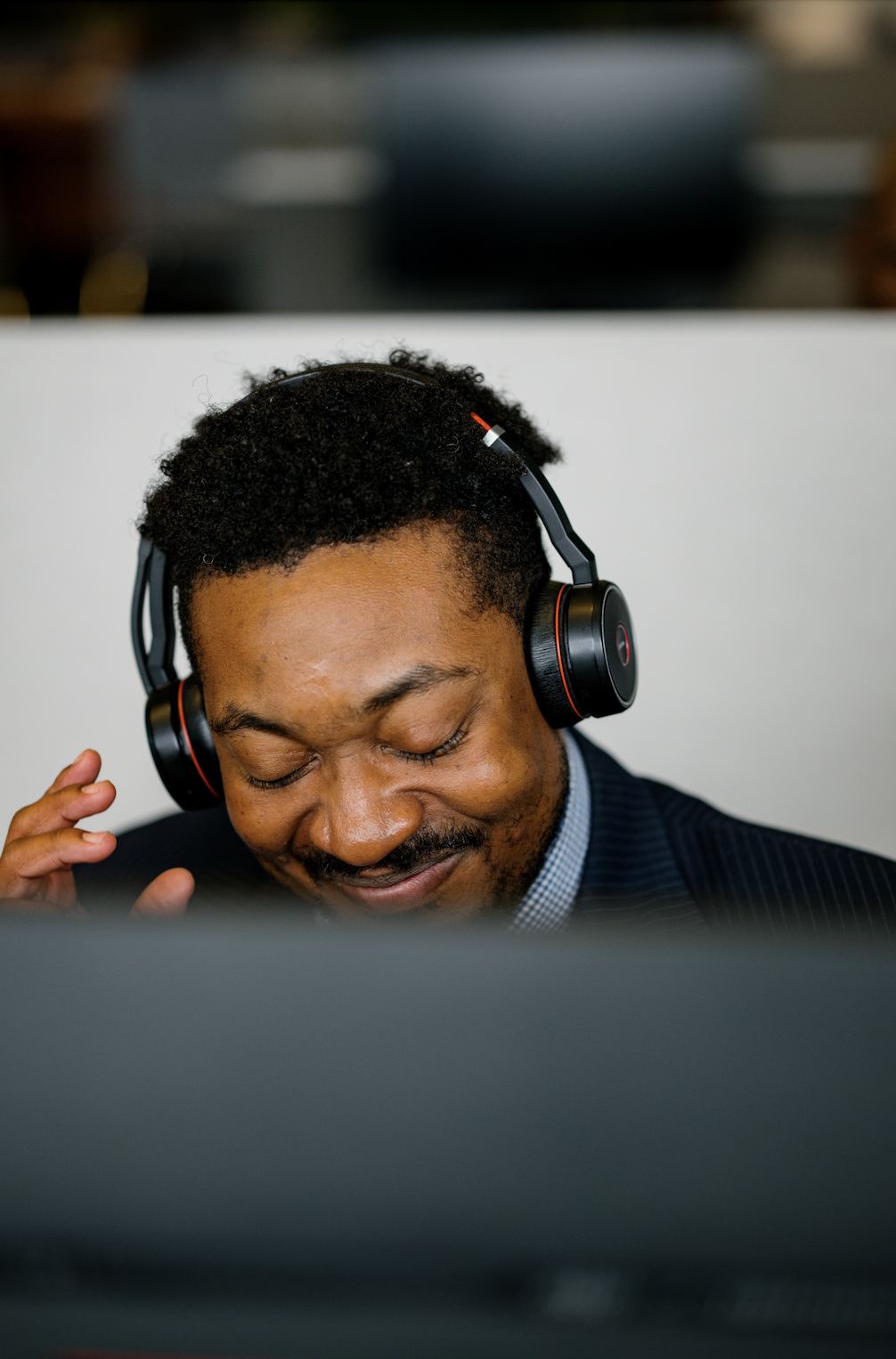 A man with curly hair and facial hair listening to music through large black headphones, smiling with his eyes closed, in an indoor setting. Detail oriented photography that highlights the small moments of a story. Corporate office, team member photo