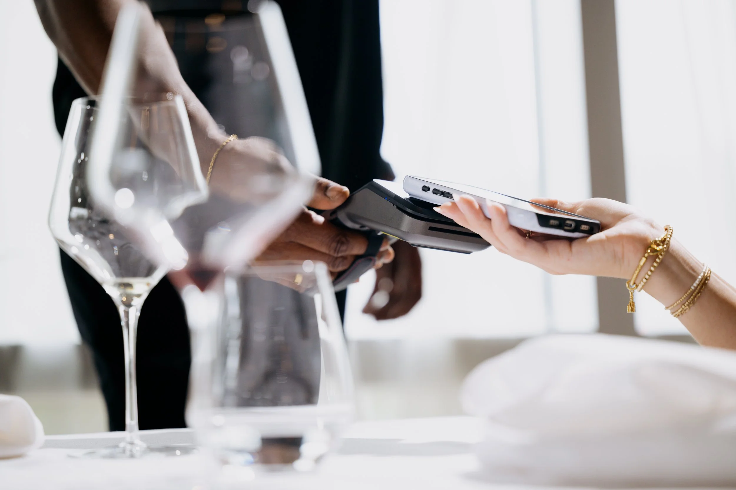 Two people exchanging a smartphone at a dining table with partially visible wine glasses and a white tablecloth.