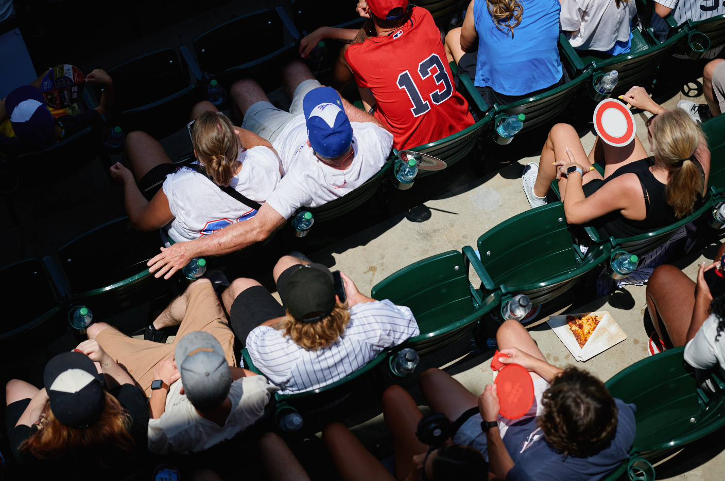 Crowd of people sitting on green stadium seats at an outdoor event, with some holding food and drinks, and sunglasses and caps visible. Sports experience photography. 