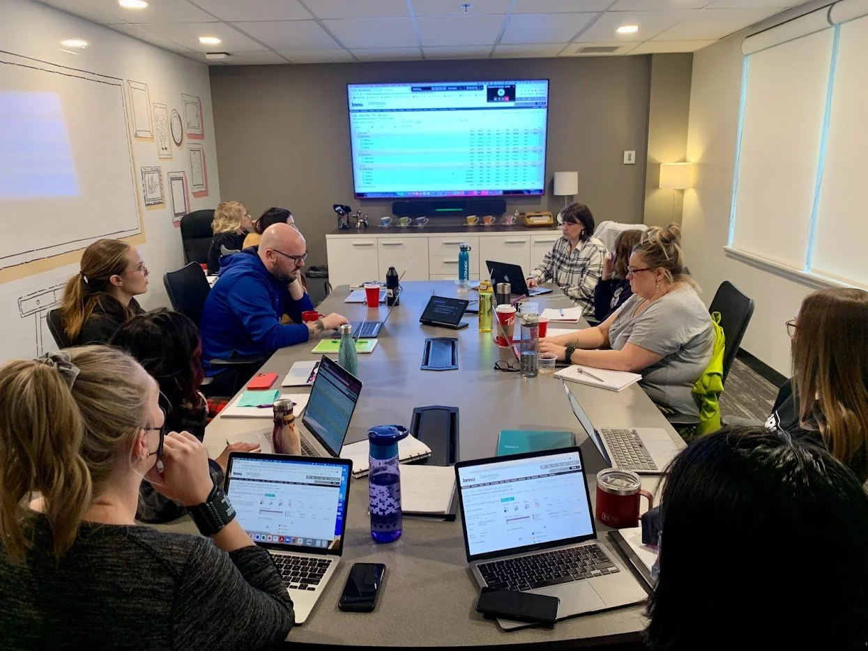 People seated around a conference table with laptops and notebooks, watching a presentation on a large screen in a modern office conference room.