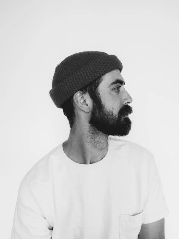 Side profile of a man with a beard and mustache, wearing a dark knit beanie and a white T-shirt, against a plain background. Photographer, creative director Grant Hindsley.
