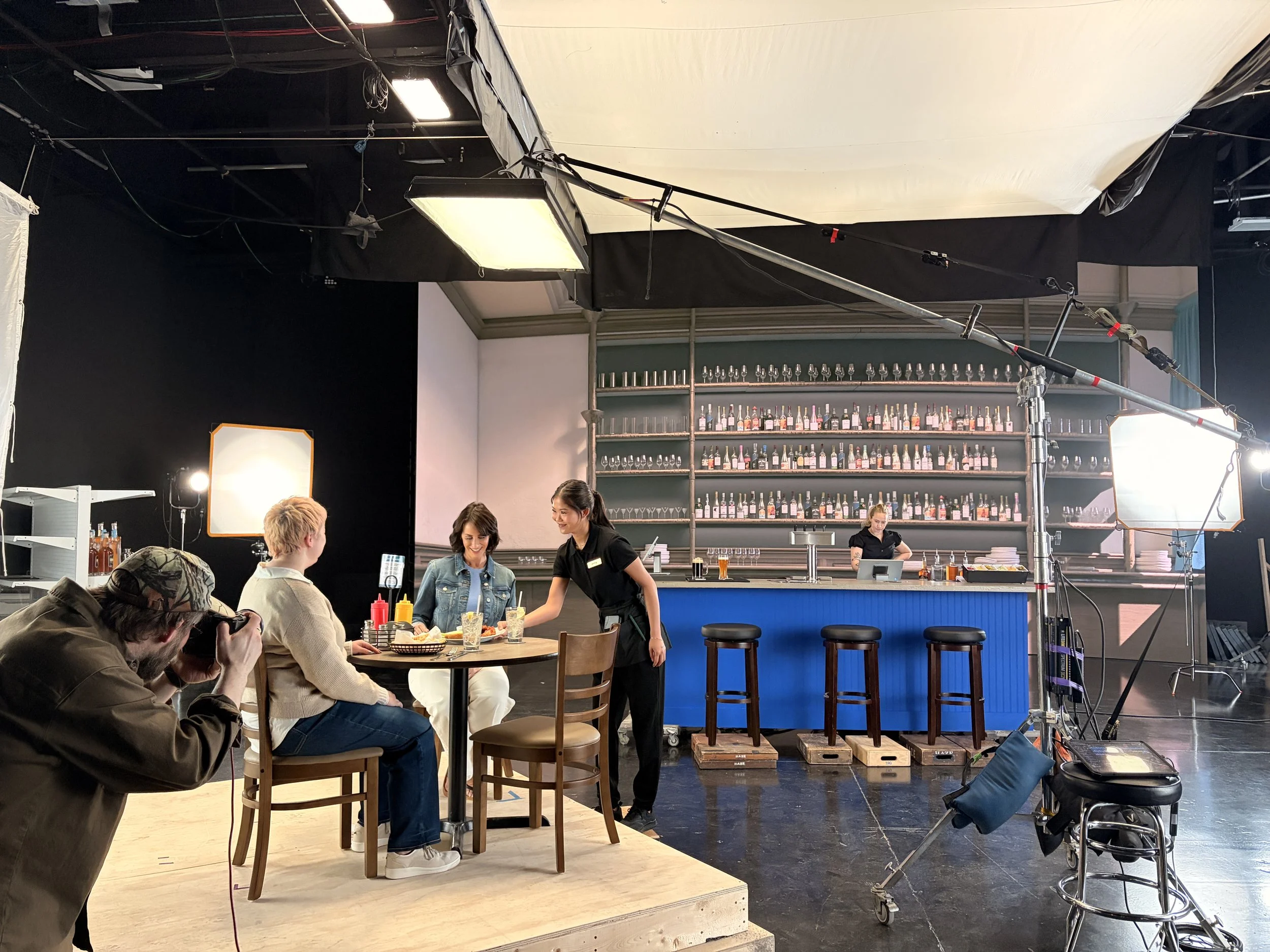 Behind-the-scenes of a film or photoshoot setup in a studio with a bar and a panel of team members, lighting equipment, and camera crew photographing a woman seated at a table.