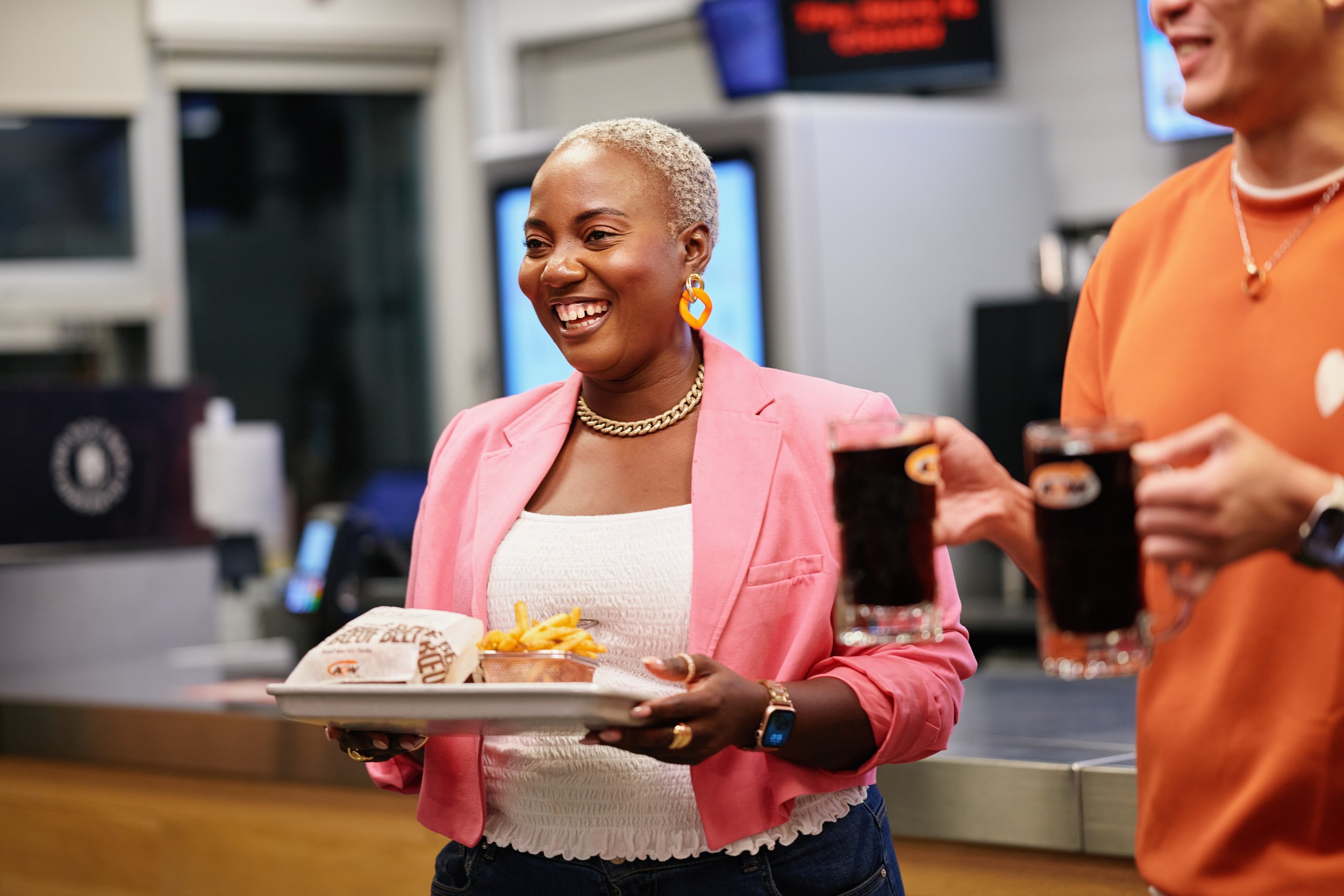 A woman with short, platinum blonde hair smiling while holding a tray with fast food, including a burger, fries, and a soda, in a casual restaurant setting. She wears a pink blazer, white top, large earrings, necklace, and a watch.