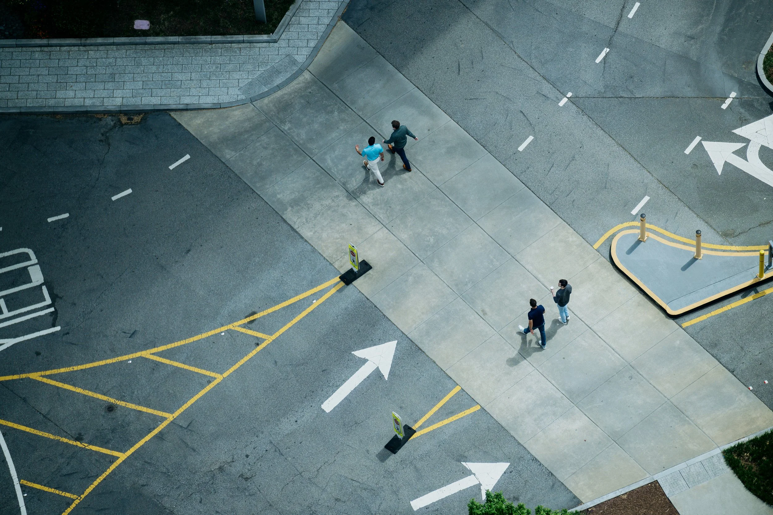 An aerial view of a city sidewalk and street, featuring pedestrians, painted parking spaces, traffic markings, and yellow and white road lines. Drone construction, city planning, photography. 