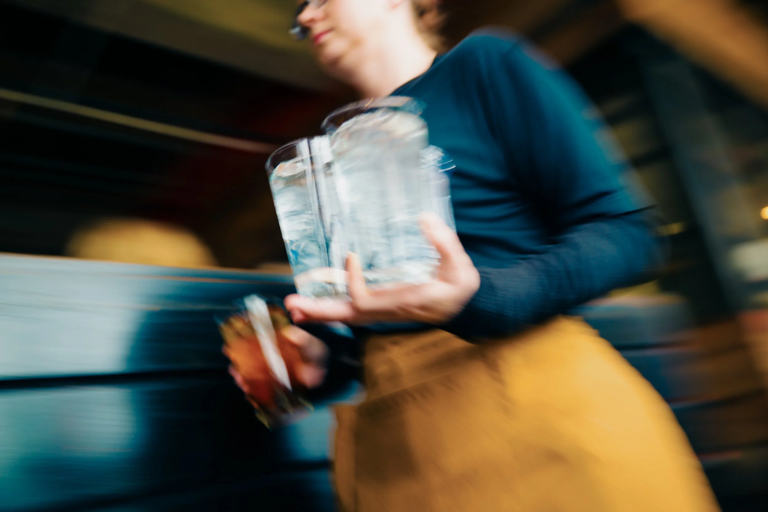 A woman with dark hair, wearing a blue jacket, is holding a large glass of water and a white plastic bag, walking through a blurred indoor space.