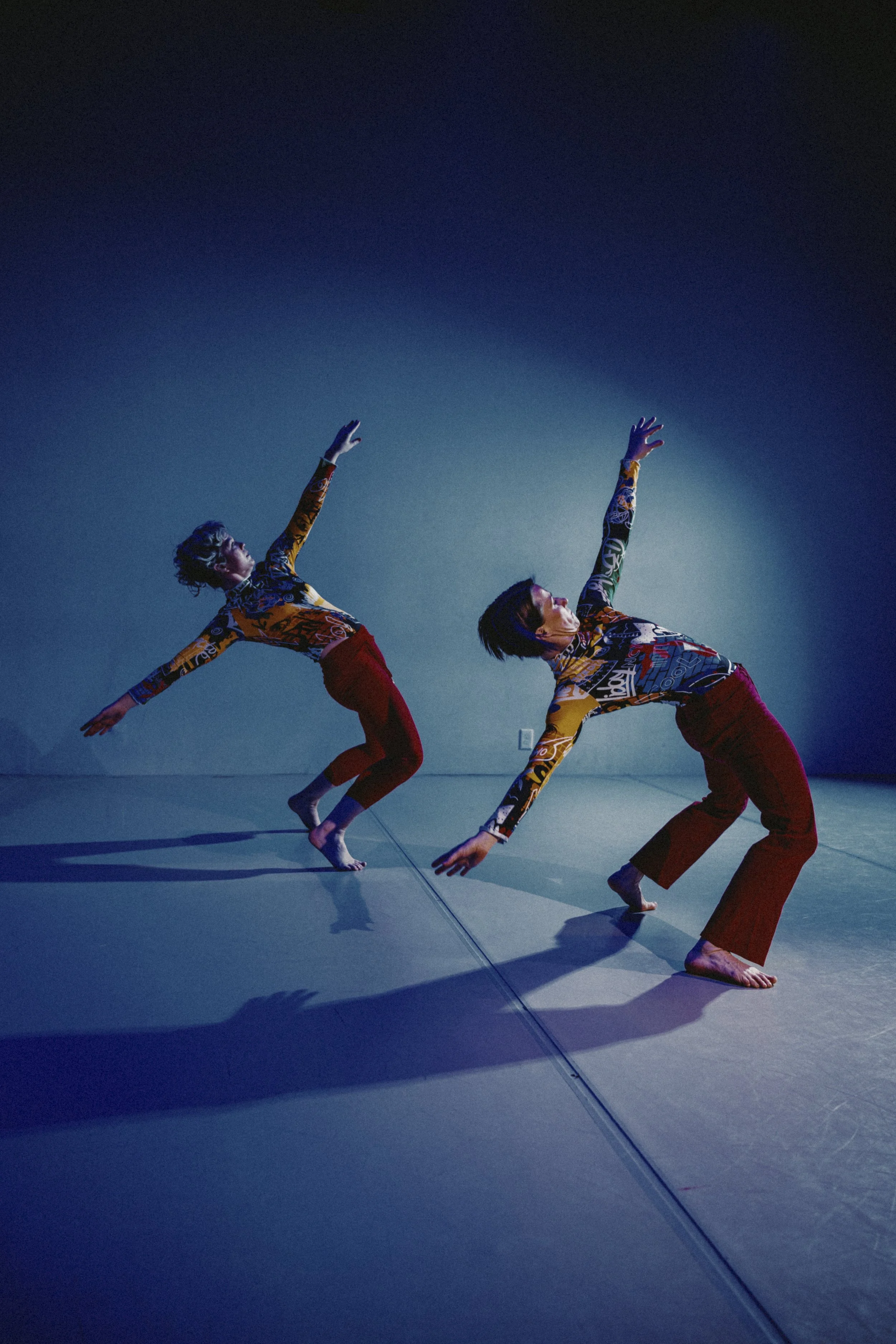 Two dancers in colorful patterned outfits perform a synchronized dance routine on a dimly lit stage, with a blue gradient background and their shadows visible on the floor.