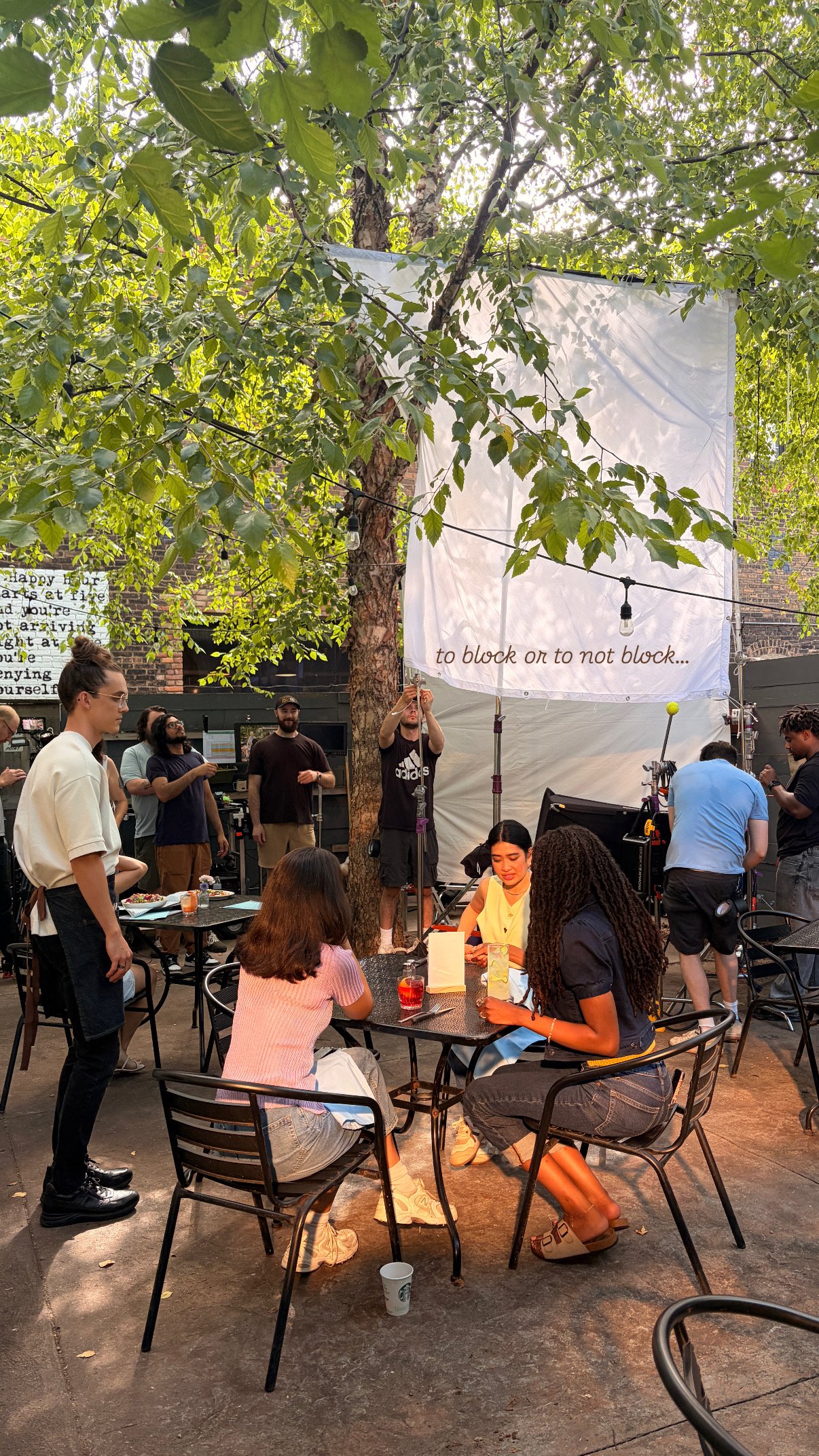 A group of people at an outdoor gathering under a tree, with some sitting at tables and others standing. There are party decorations including a large white banner with the words "to block or to not block..." and string lights hanging from the branches.