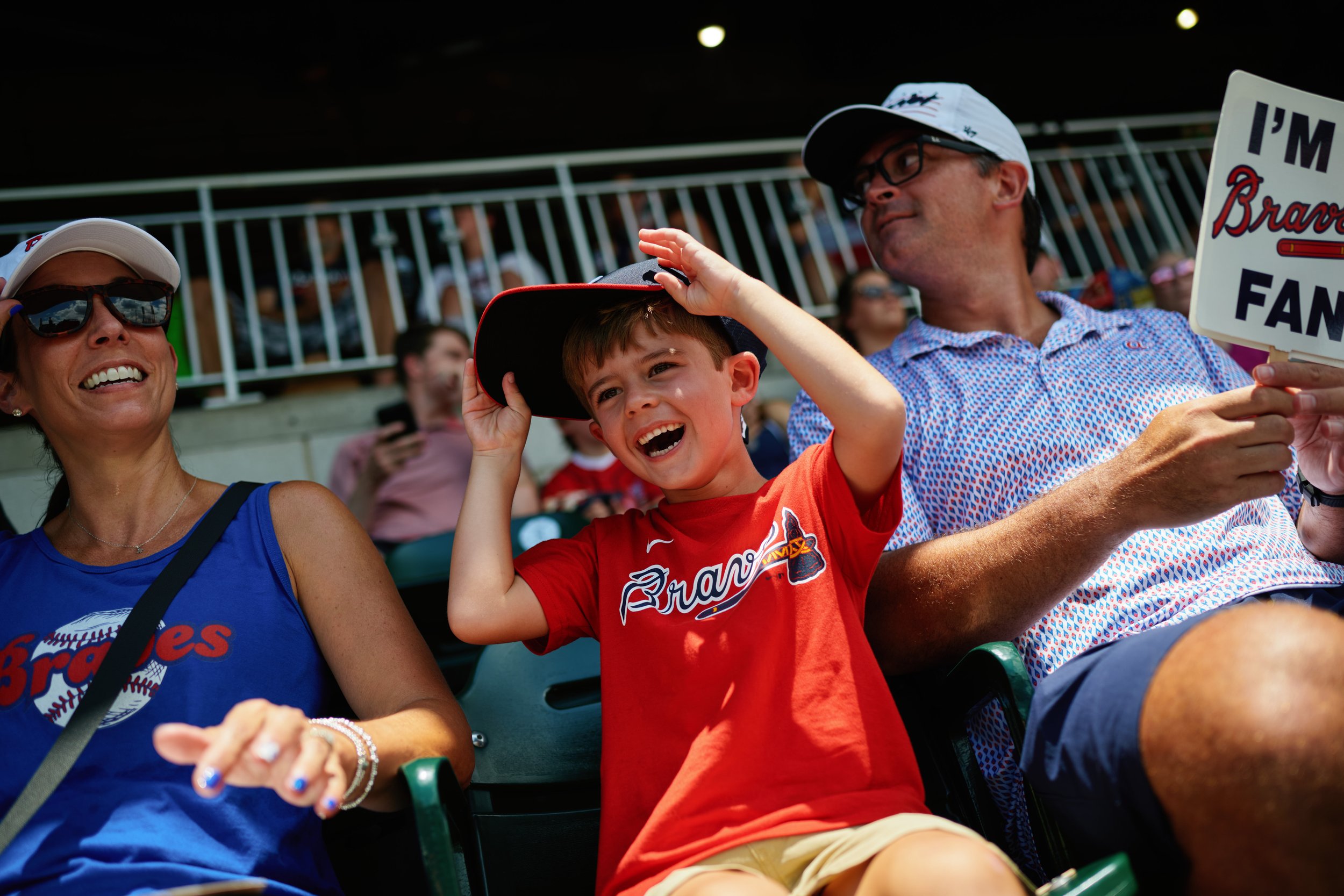 People attending a sports event, including a woman wearing sunglasses and a blue shirt, a young boy in a red shirt with a baseball cap, and a man holding a sign that says "I'M Braves FAN." all sitting in stadium seats.