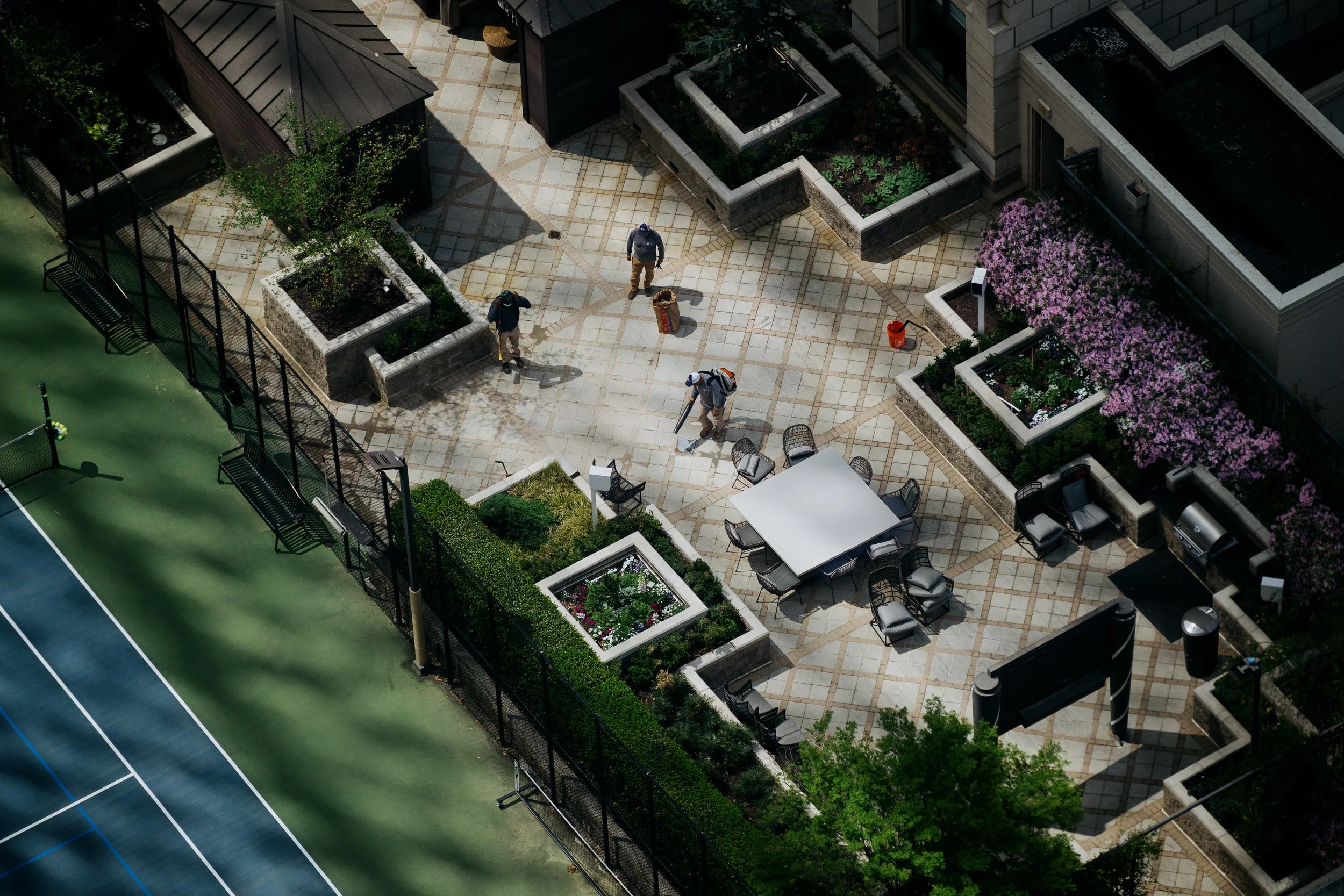 An aerial view of a rooftop terrace with potted plants, trees, seating areas, and a concrete walkway, adjacent to a tennis court with green surface and blue lines.