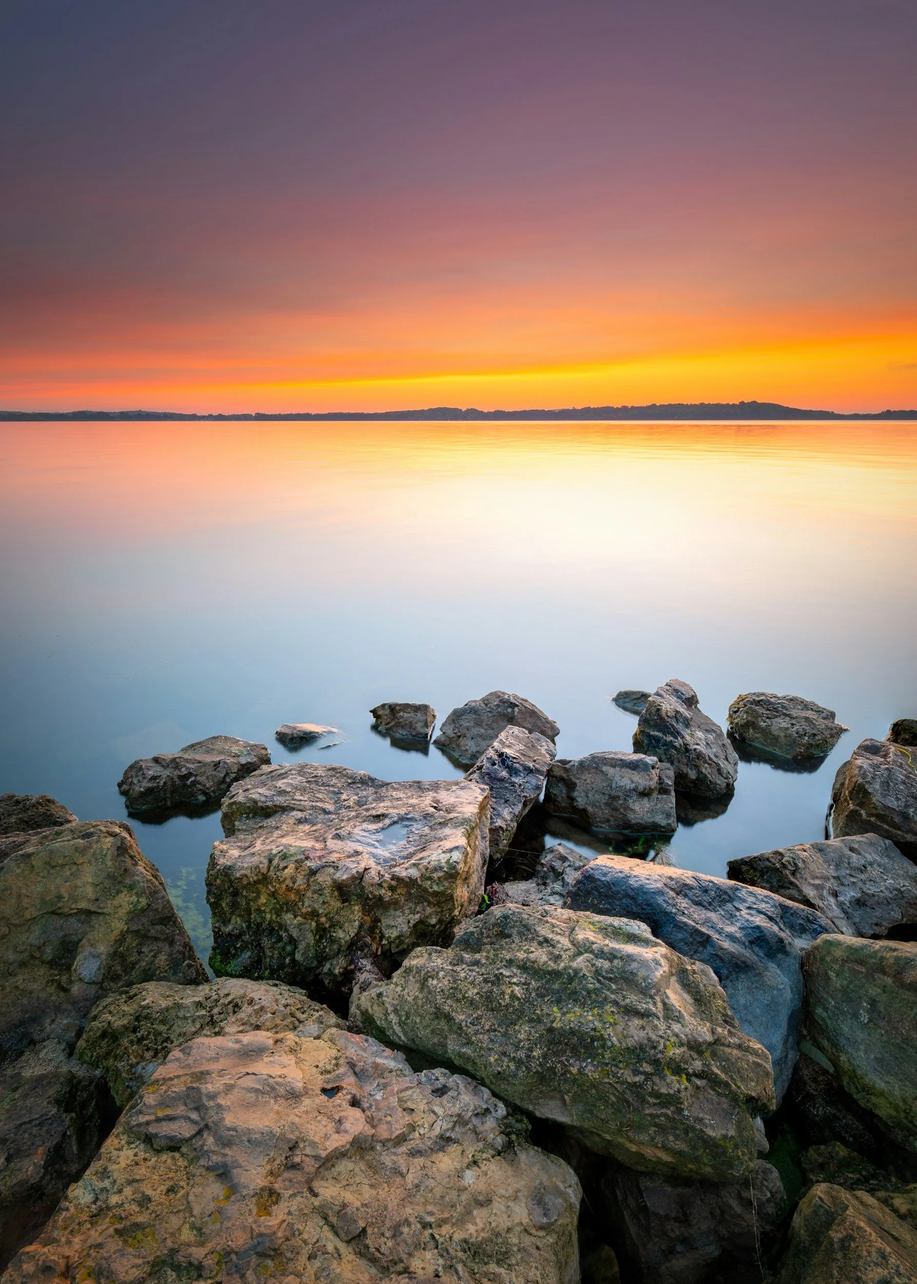 Rocks along a lake shore during sunset with a colorful sky reflected on the water.
