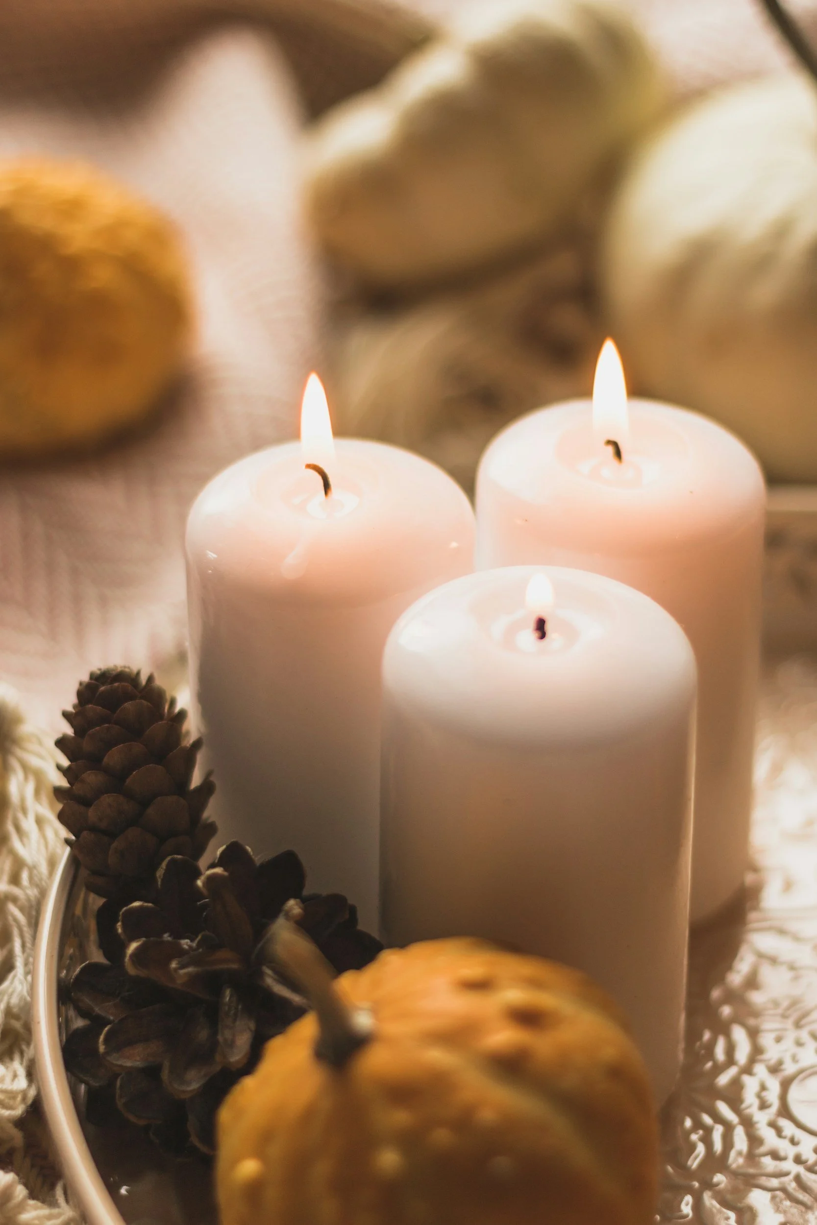 Three lit white candles surrounded by pinecones, gourds, and decorative items on a reflective surface.