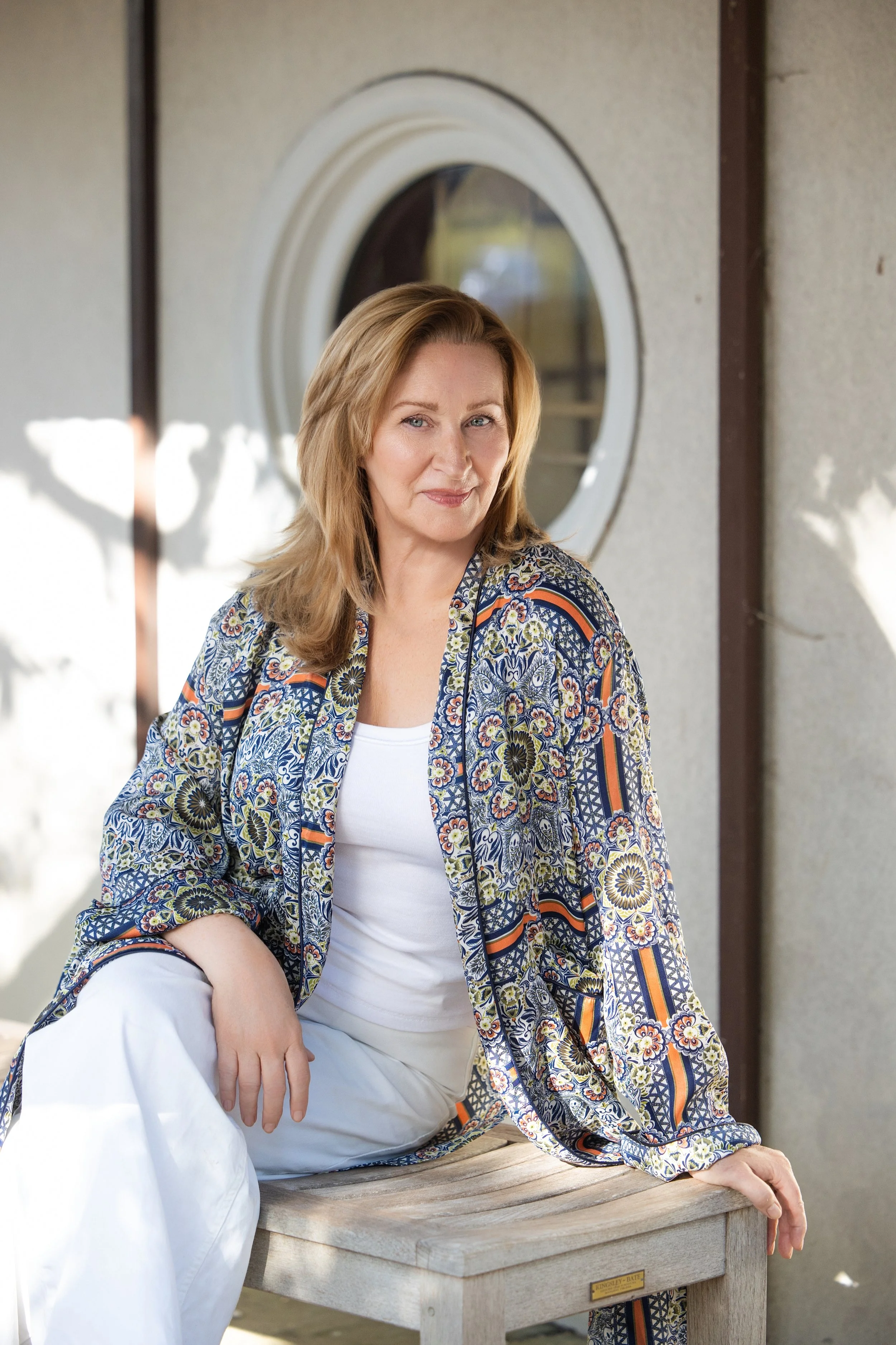 A woman with blonde hair sitting outdoors on a wooden bench next to a building with a round window. She is wearing a patterned jacket and white pants, smiling softly.