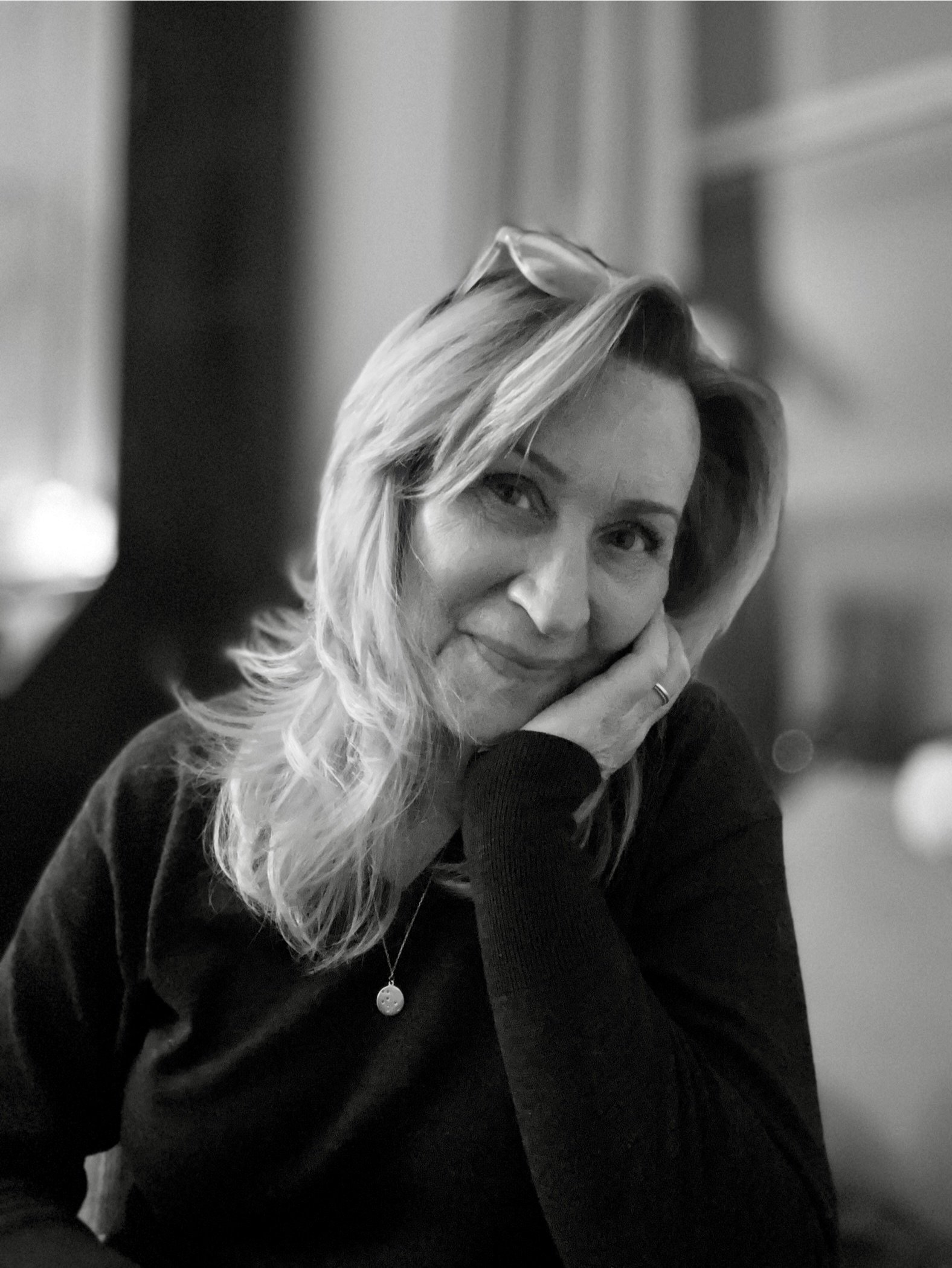 Black and white portrait of a woman with shoulder-length hair, resting her face on her hand, wearing glasses on her head, a necklace, and a dark sweater.