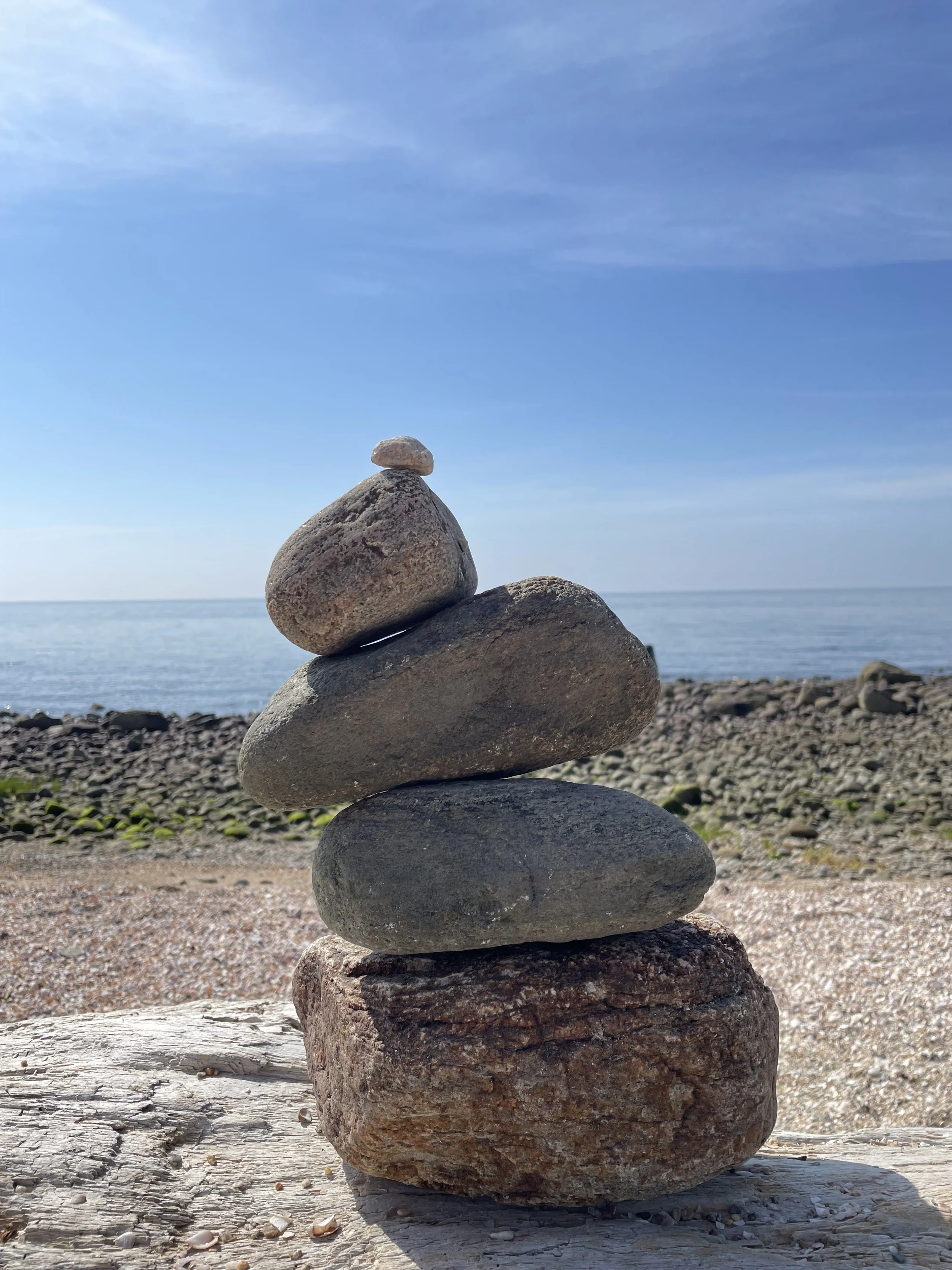 Stacked rocks on a beach with a shoreline and ocean in the background under a blue sky.