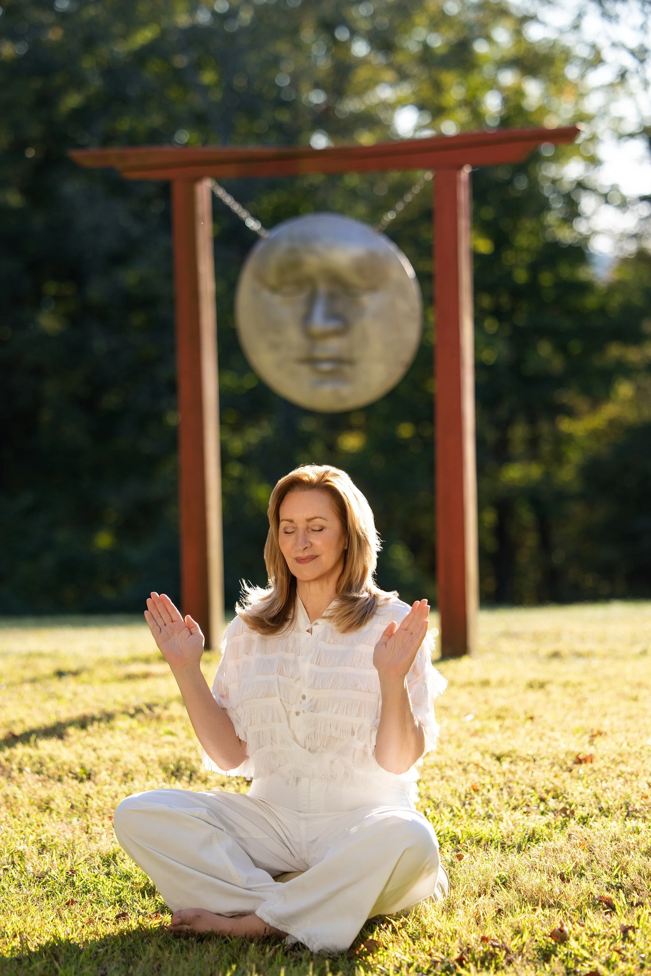 A woman practicing yoga or meditation outdoors in a park, sitting cross-legged on the grass with eyes closed, in front of a large moon face sculpture hanging from a wooden frame.