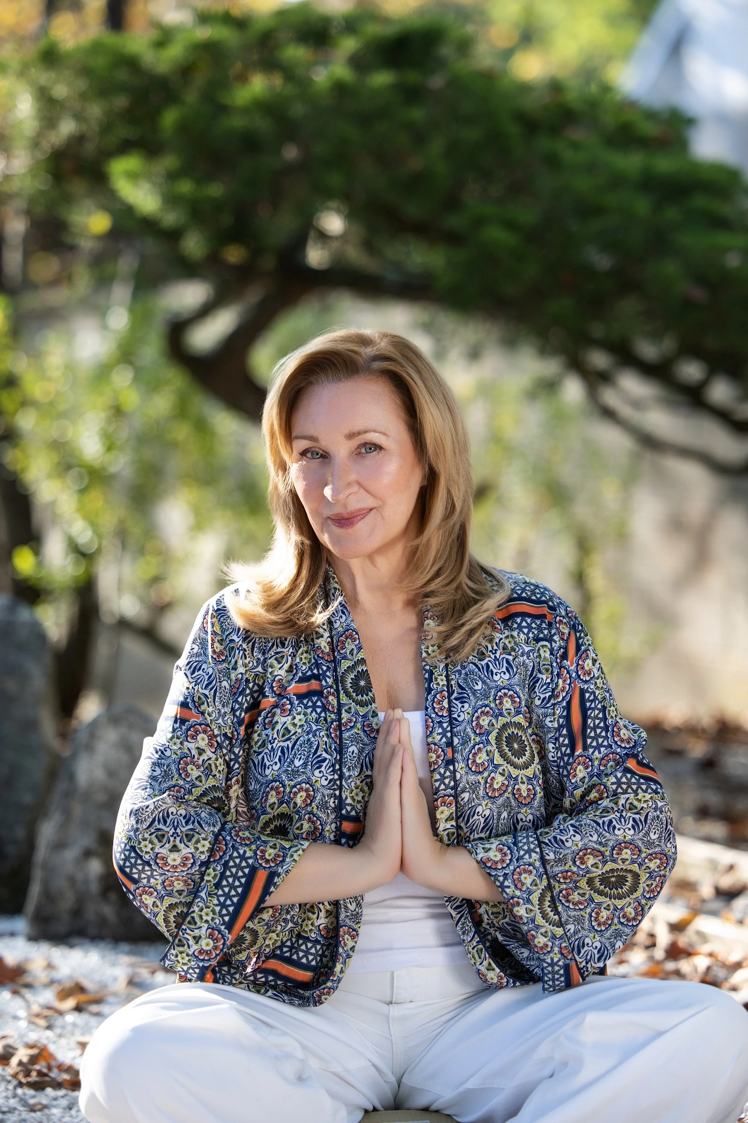 A woman with blonde hair practicing yoga outdoors, sitting cross-legged with hands together in a prayer position, surrounded by trees and rocks.