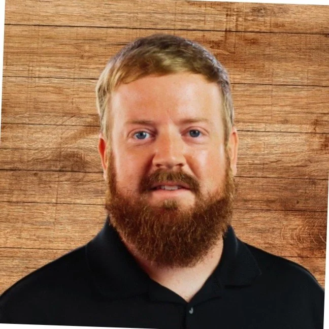 A man with a red beard, blue eyes, and short blonde hair, wearing a black collared shirt, standing against a wooden wall background.