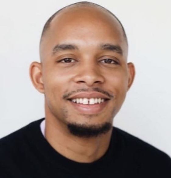 A smiling man with a shaved head and goatee in front of a plain light background.