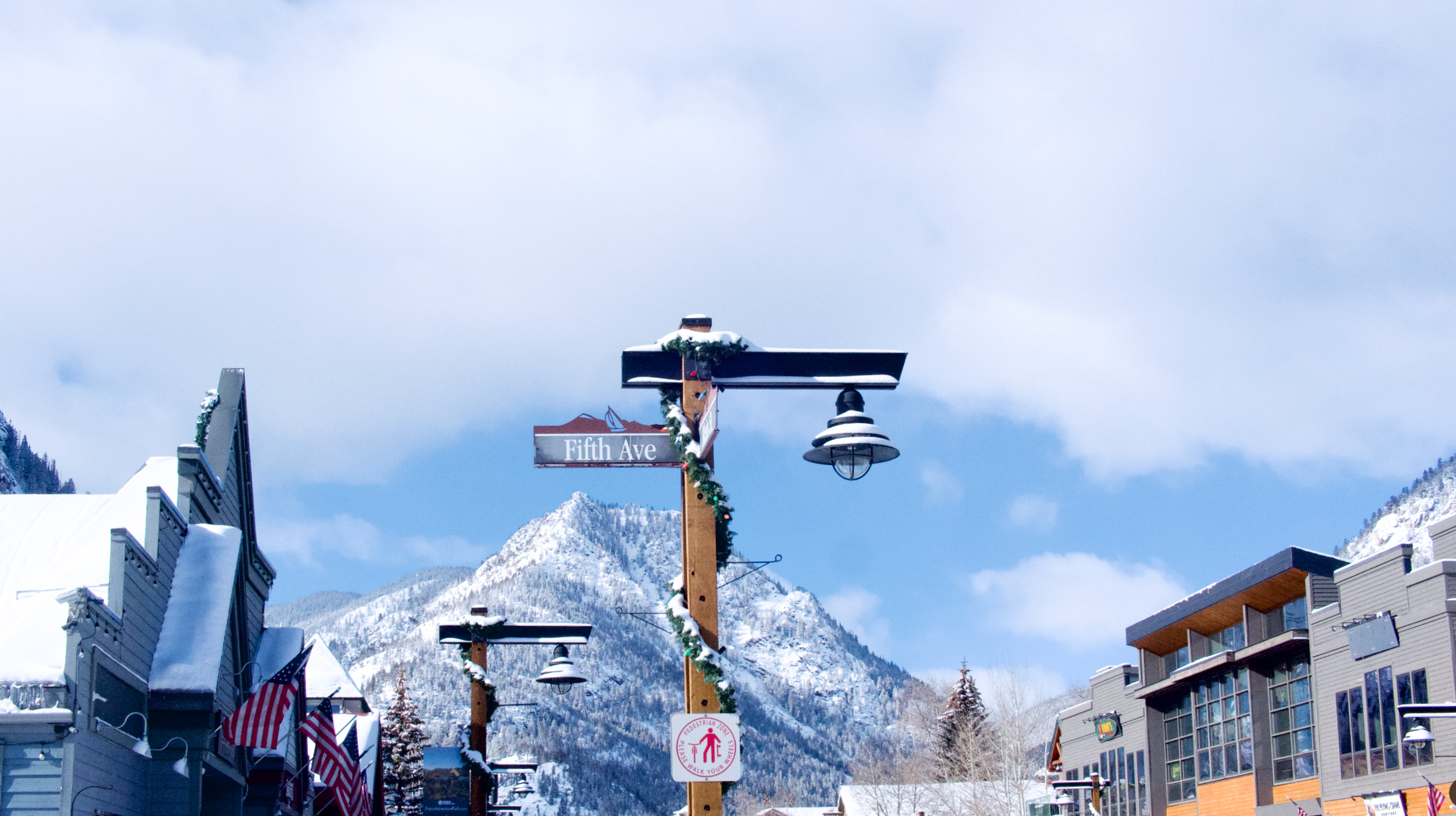 Snow-covered street scene in a mountain town with buildings, street signs, decorated light poles, American flags, and a mountainous backdrop under a partly cloudy sky.