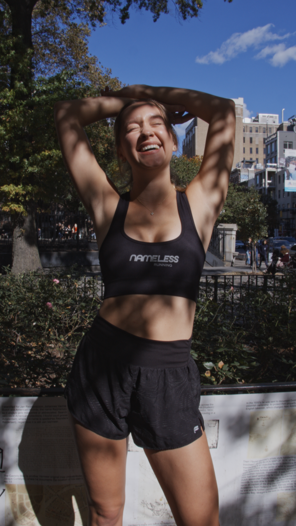 Young woman smiling with arms raised behind her head outdoors in a park, wearing a black sports bra and shorts, with trees, buildings, and a blue sky in the background.