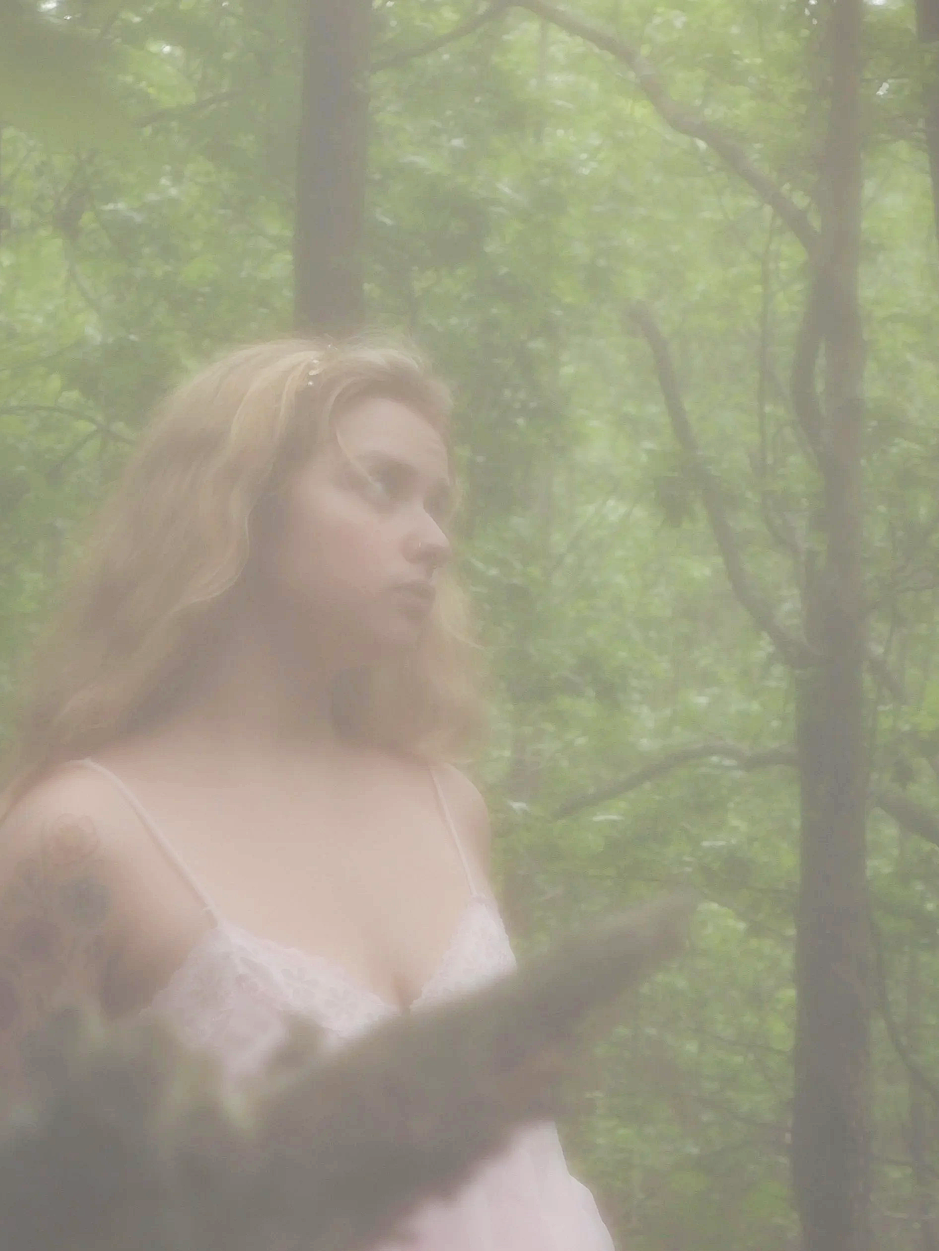 A woman with long, light brown hair wearing a white dress with lace, standing in a forest with green leaves and trees, as seen through a foggy or misty window.