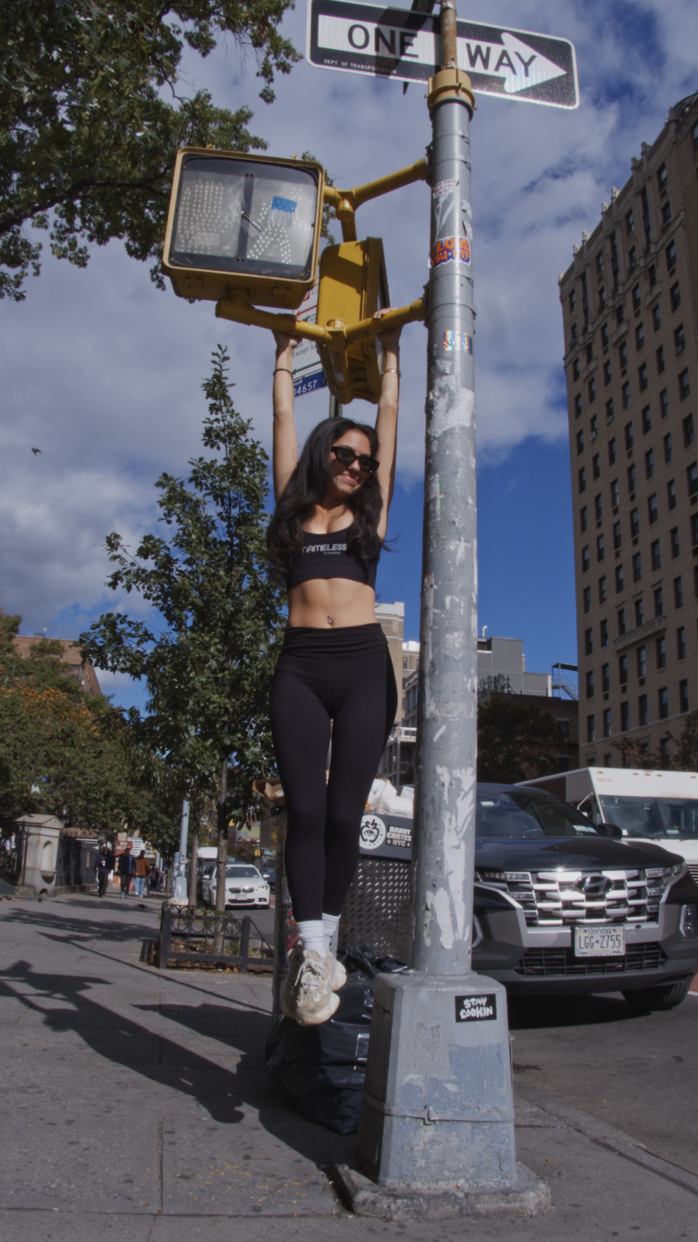 A woman hanging from a streetlight pole with her arms, smiling, wearing sunglasses, a black crop top, black leggings, and white sneakers, on a city sidewalk with cars and buildings in the background under a partly cloudy sky.